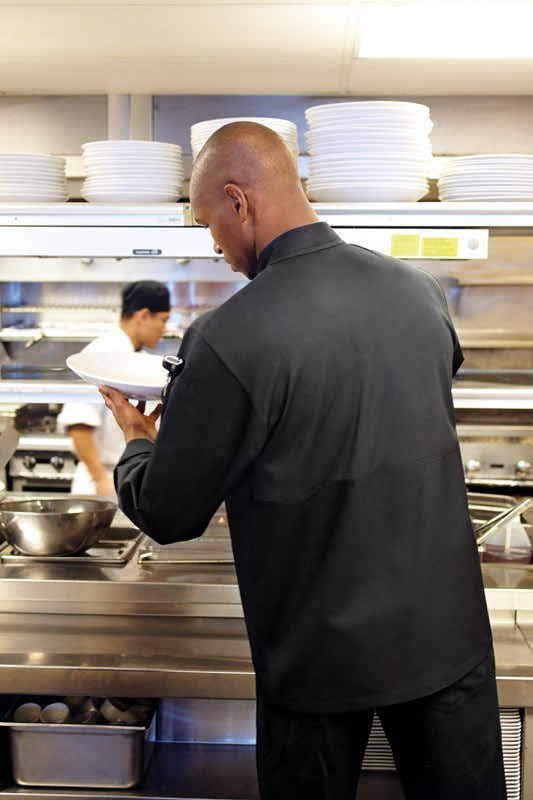 A black men's chef jacket is being worn in a kitchen setting, with another chef visible in the background.