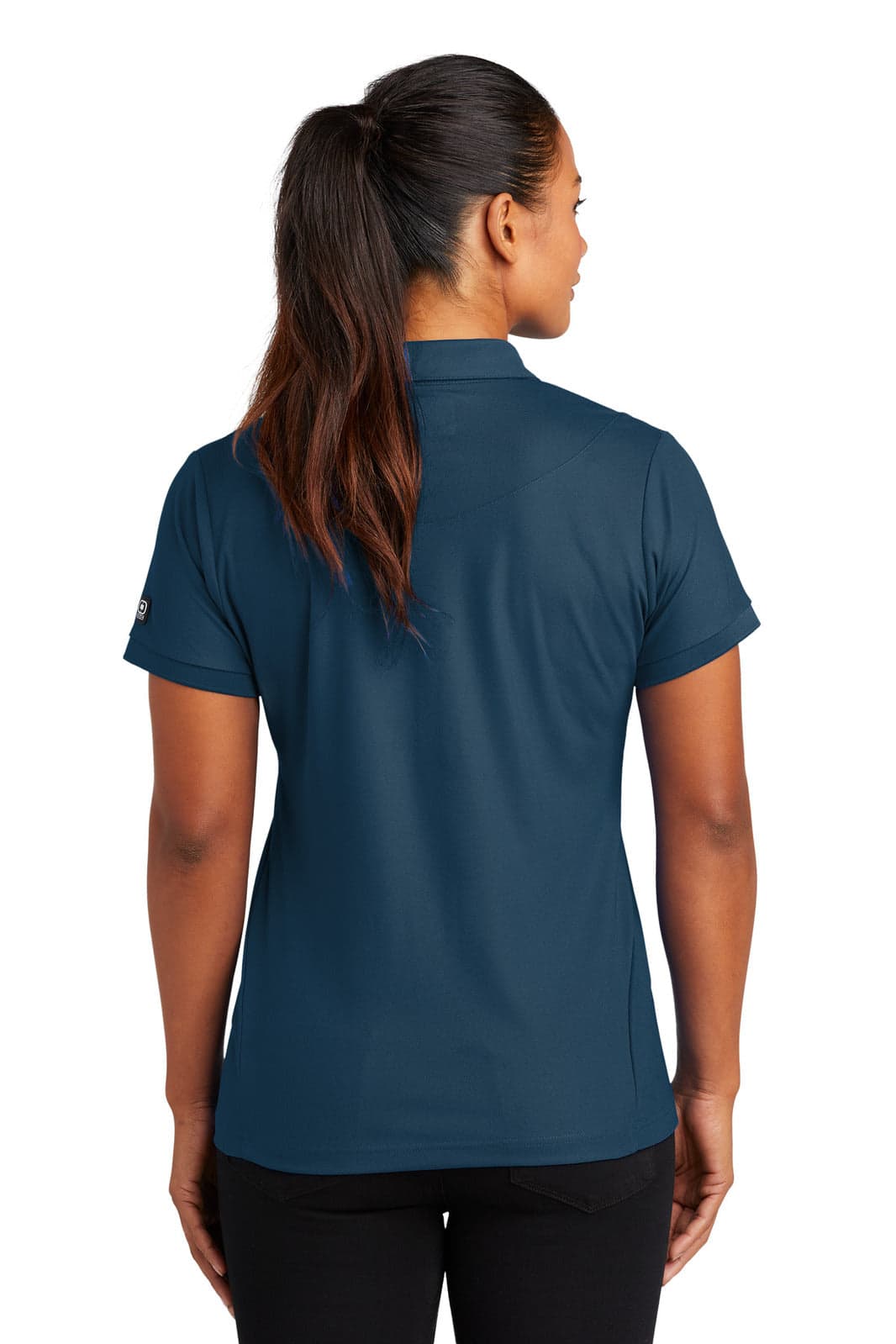 A woman is wearing a dark blue OGIO Jewel Polo shirt, viewed from the back. The shirt has short sleeves and a branded logo.
