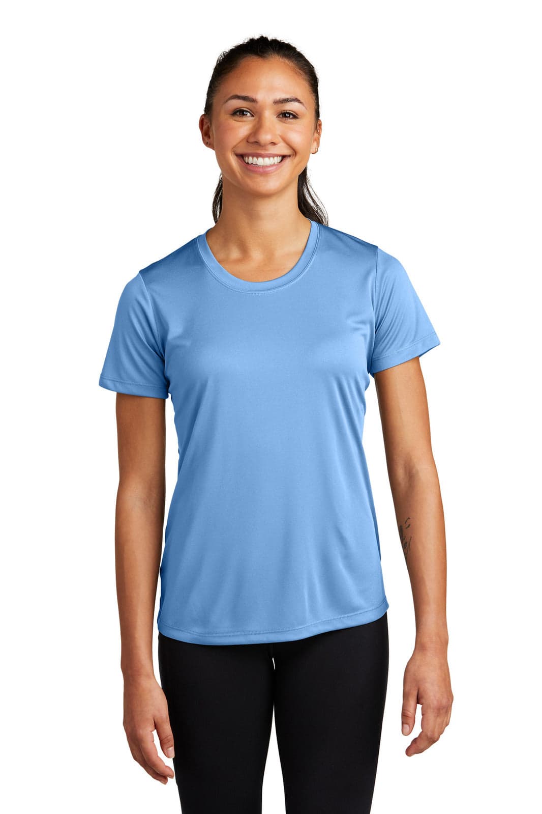 A woman in a blue athletic t-shirt stands smiling, featuring short sleeves and a comfortable fit. The shirt has a logo.