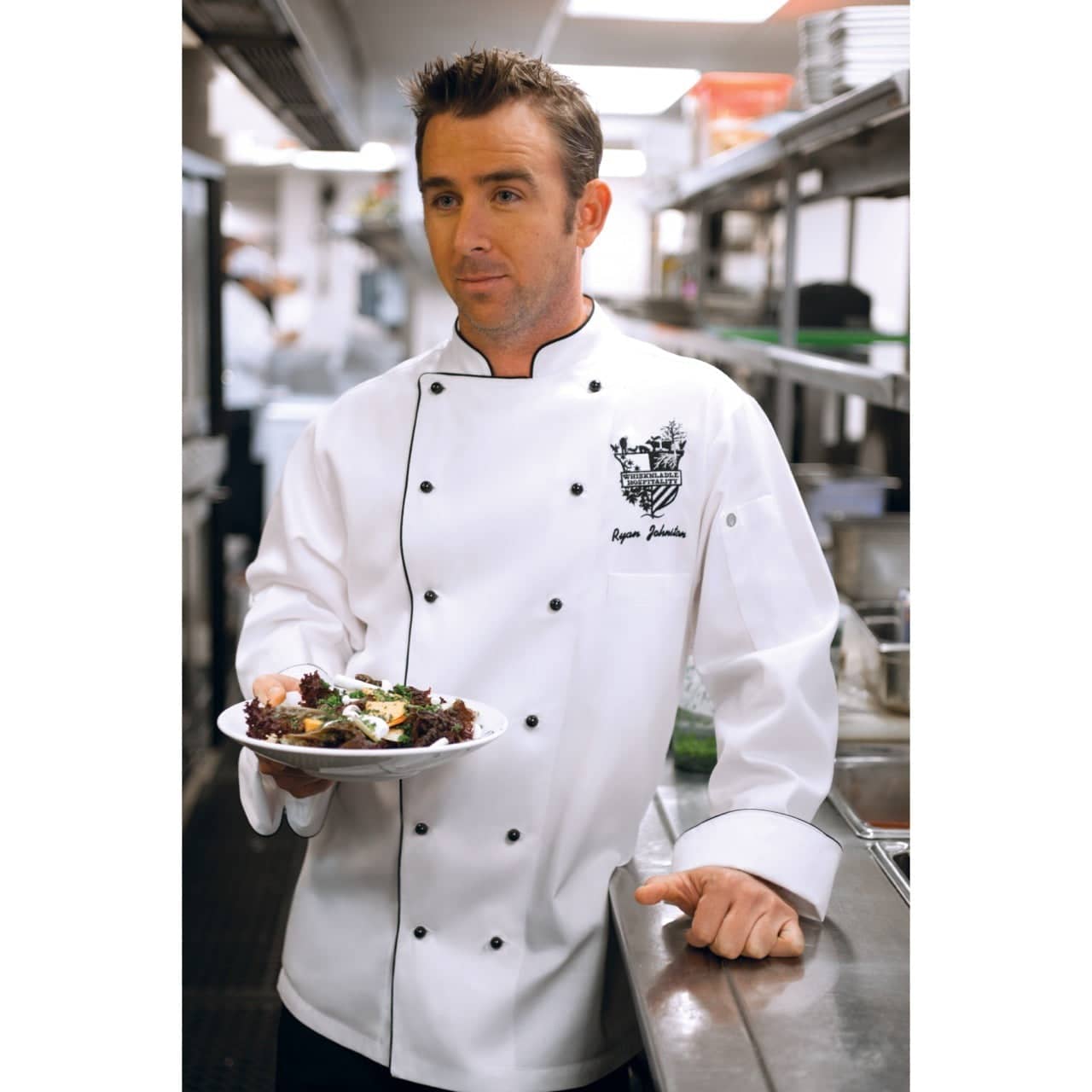 A chef jacket in white, featuring black buttons and a logo, worn by a man holding a plate of food in a kitchen setting.