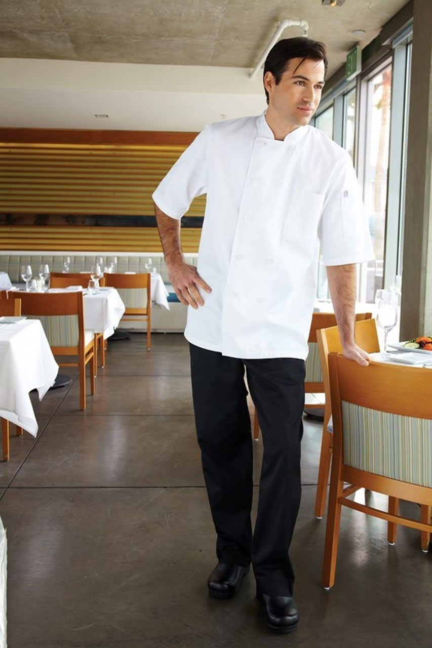 A man in a white chef jacket and black baggy chef pants stands in a restaurant setting, with tables and chairs in view.