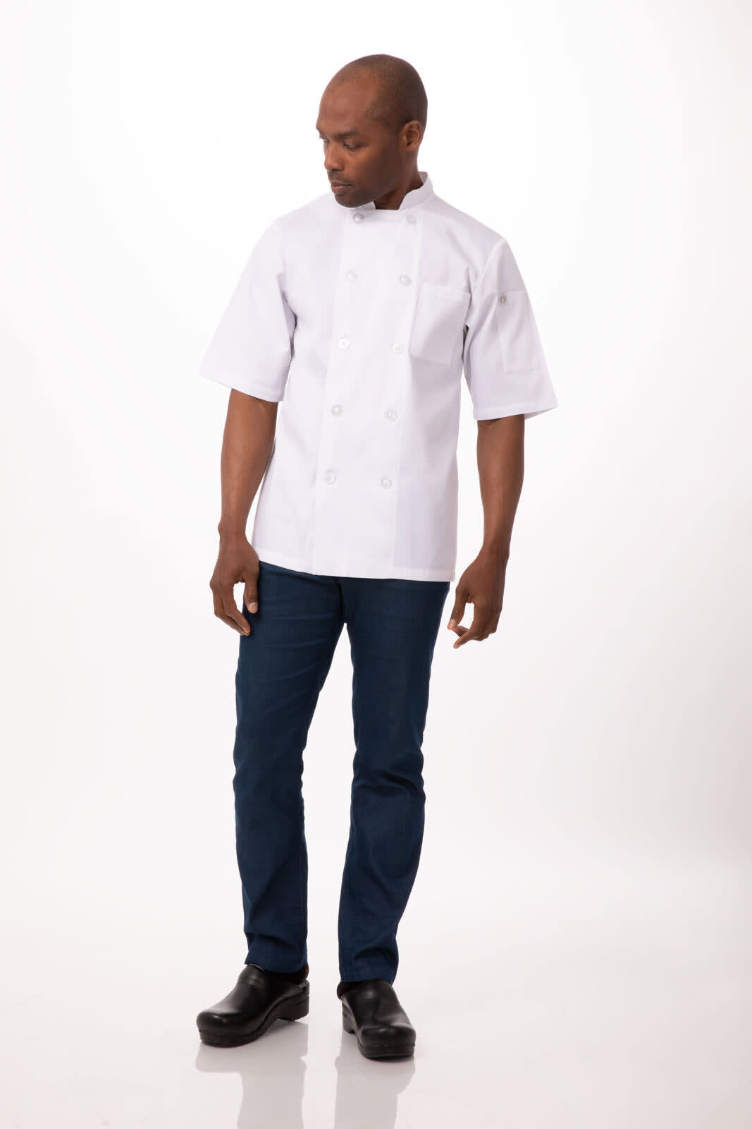 A man wearing a white chef jacket and dark blue pants, standing in a neutral setting. The jacket features a logo.