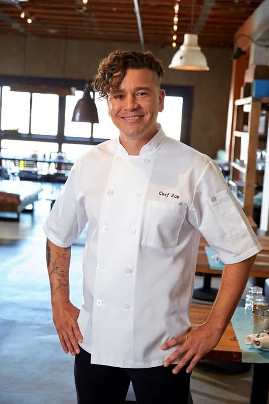 A man wearing a white chef jacket smiles in a modern restaurant setting, showcasing a professional look.