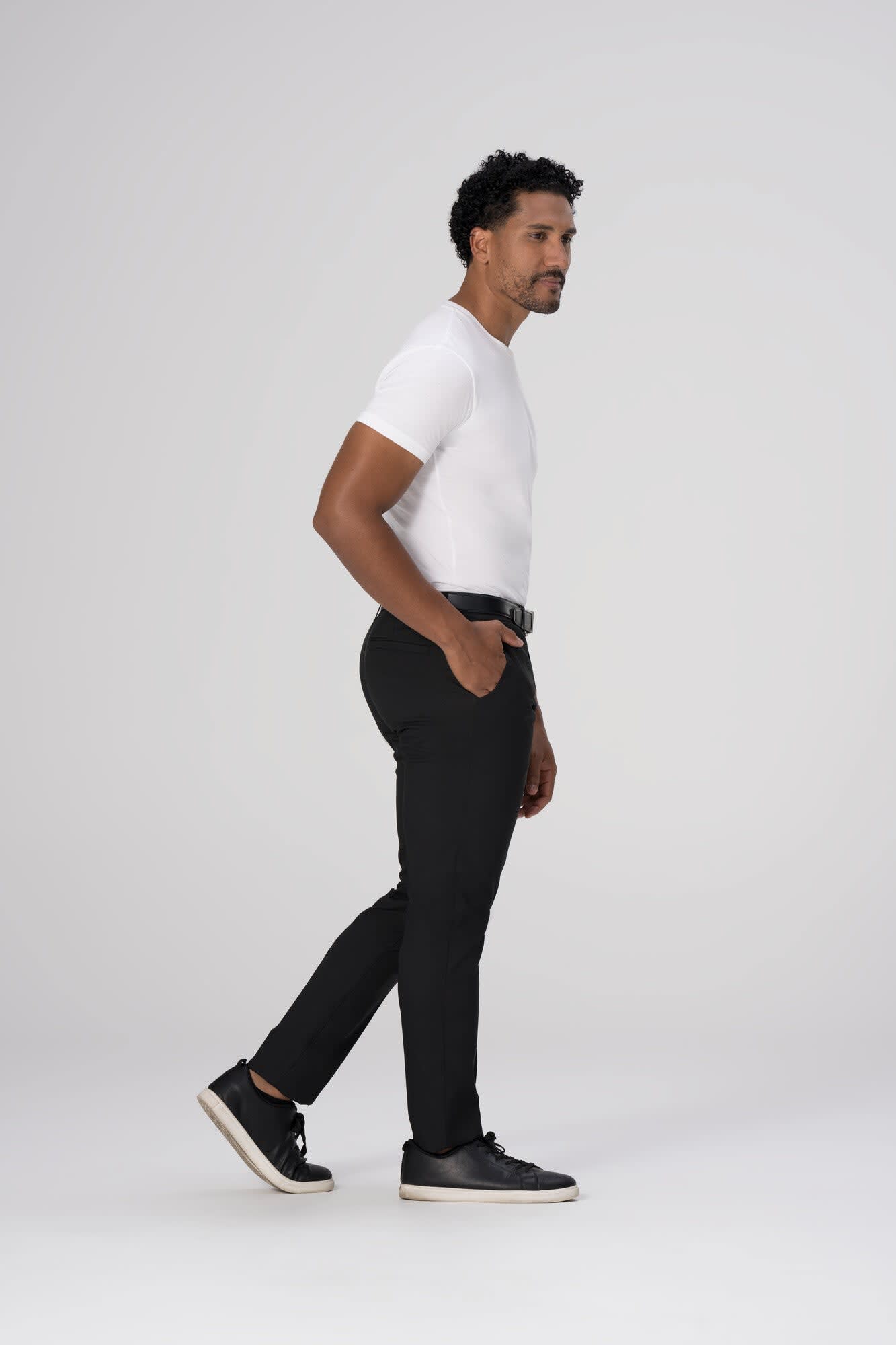 A man stands wearing black poly stretch pants and a fitted white t-shirt, with a sleek belt and casual shoes.