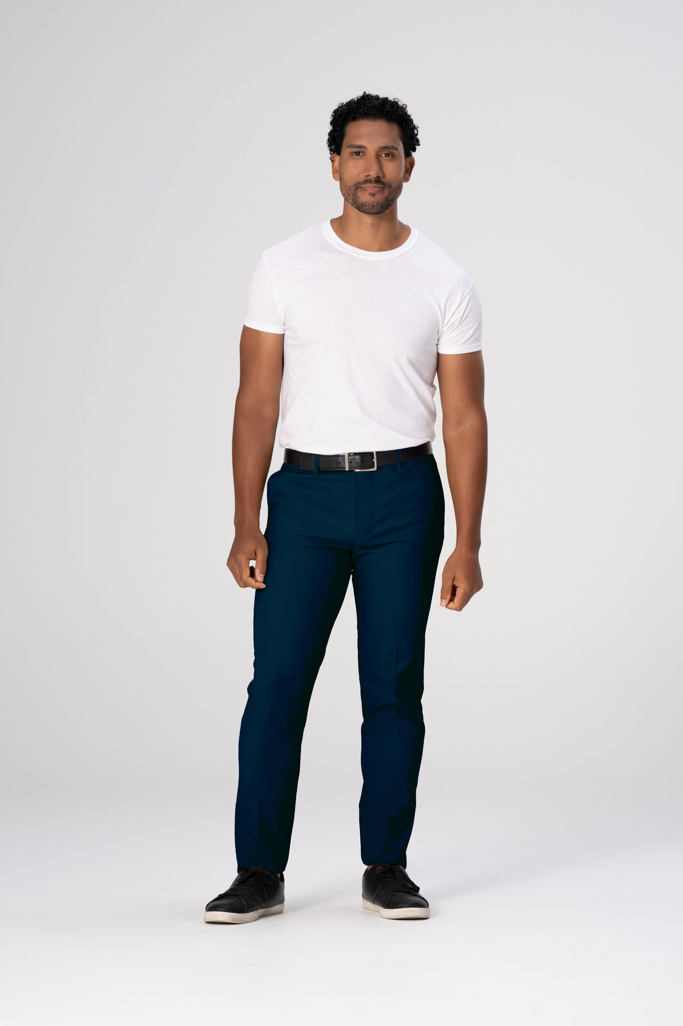 A man stands wearing navy poly stretch pants and a white t-shirt, paired with black shoes. The image has a plain background.