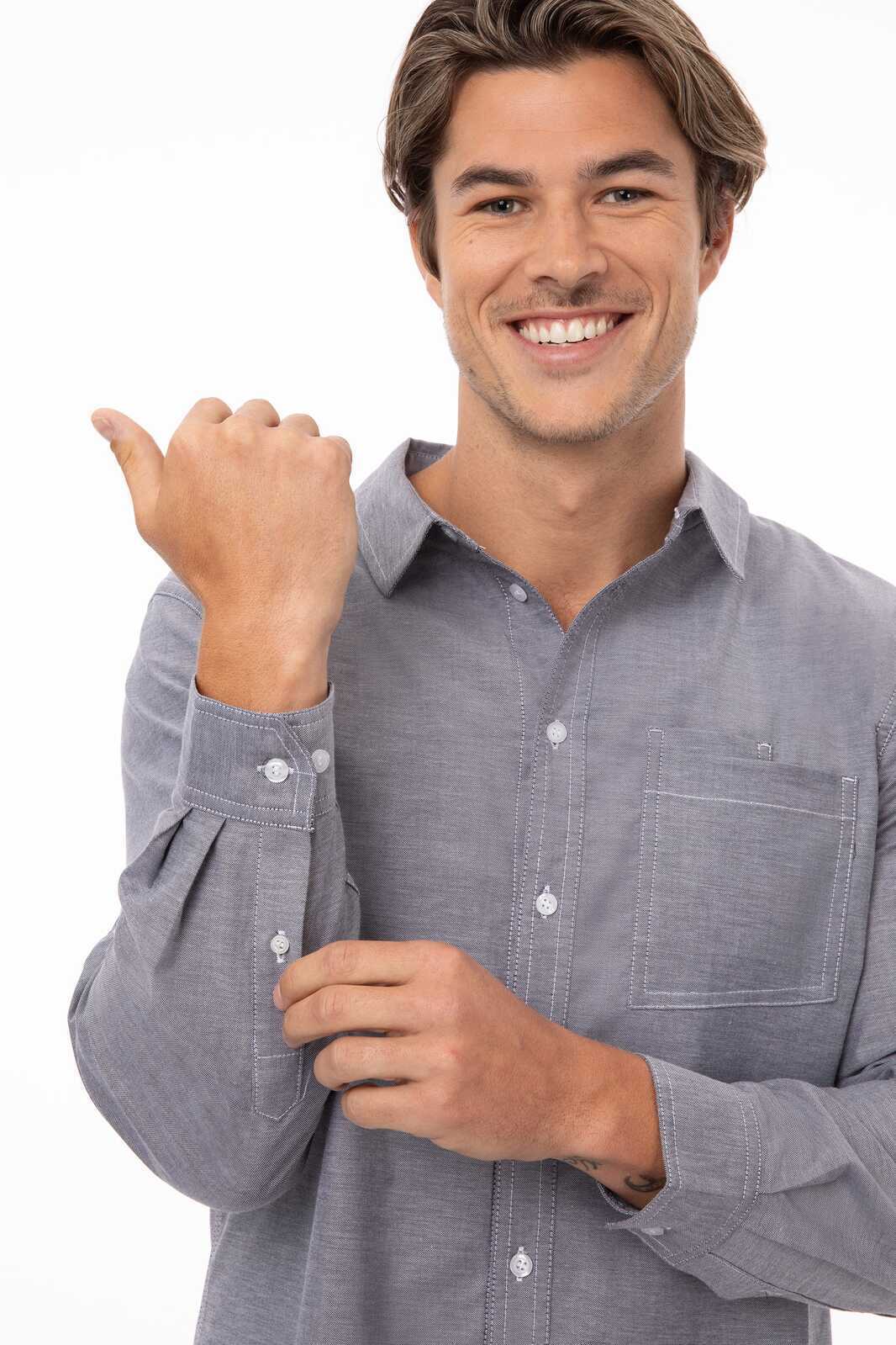 A man in a blue button-up shirt is smiling and adjusting the cuff of his shirt. The shirt features a pocket and a logo.