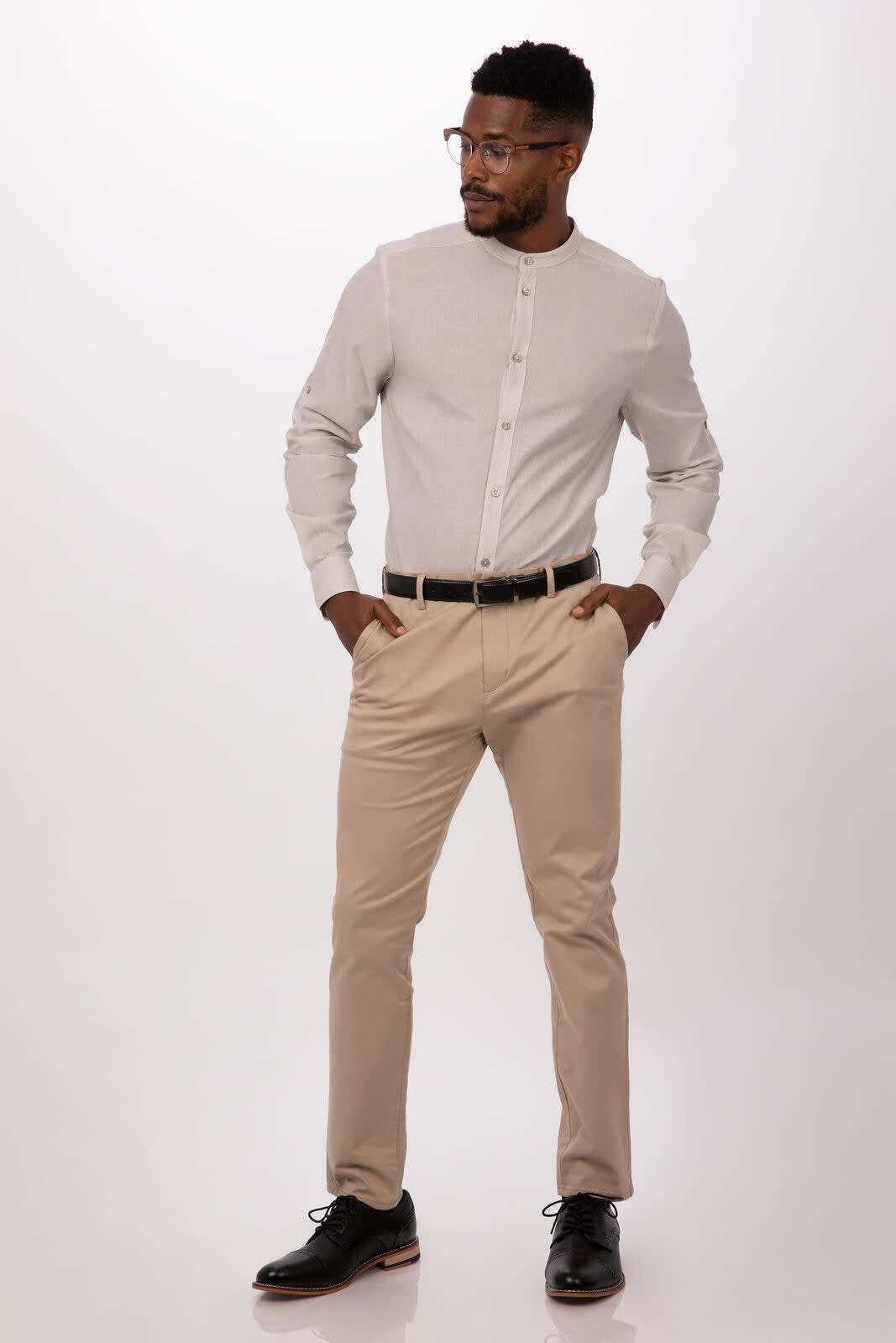 A man in a grey button-up shirt and beige trousers, standing confidently with hands in pockets. The shirt features a logo.
