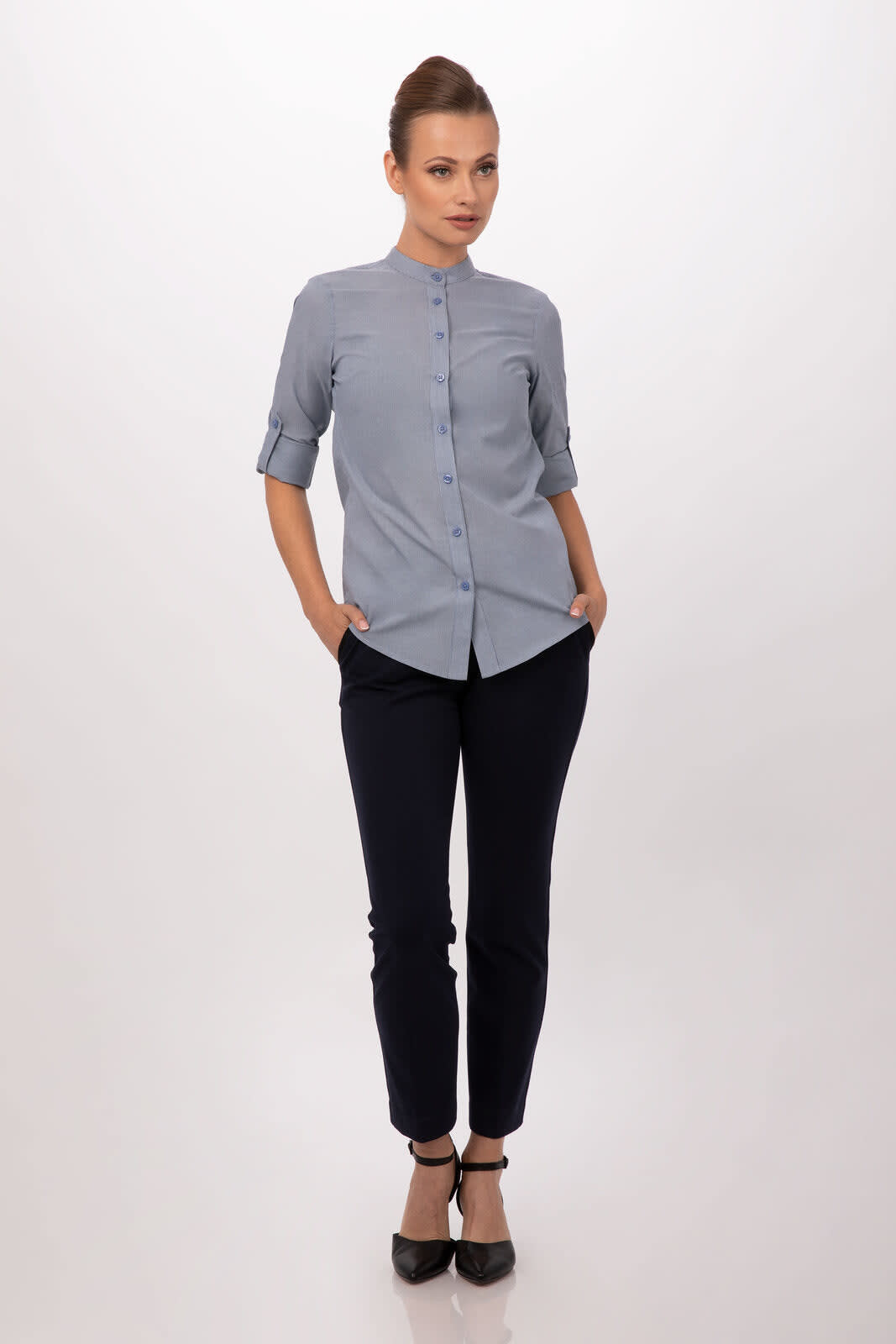 A woman models a light blue button-up shirt paired with dark trousers and heels, featuring a logo.