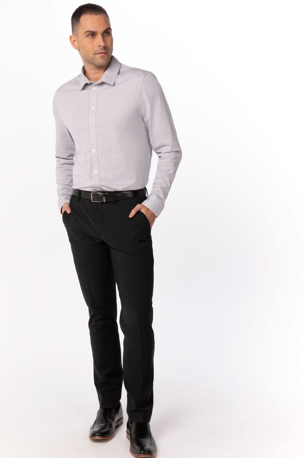 A man in a light grey button-up shirt and black trousers stands confidently with his hands in his pockets.