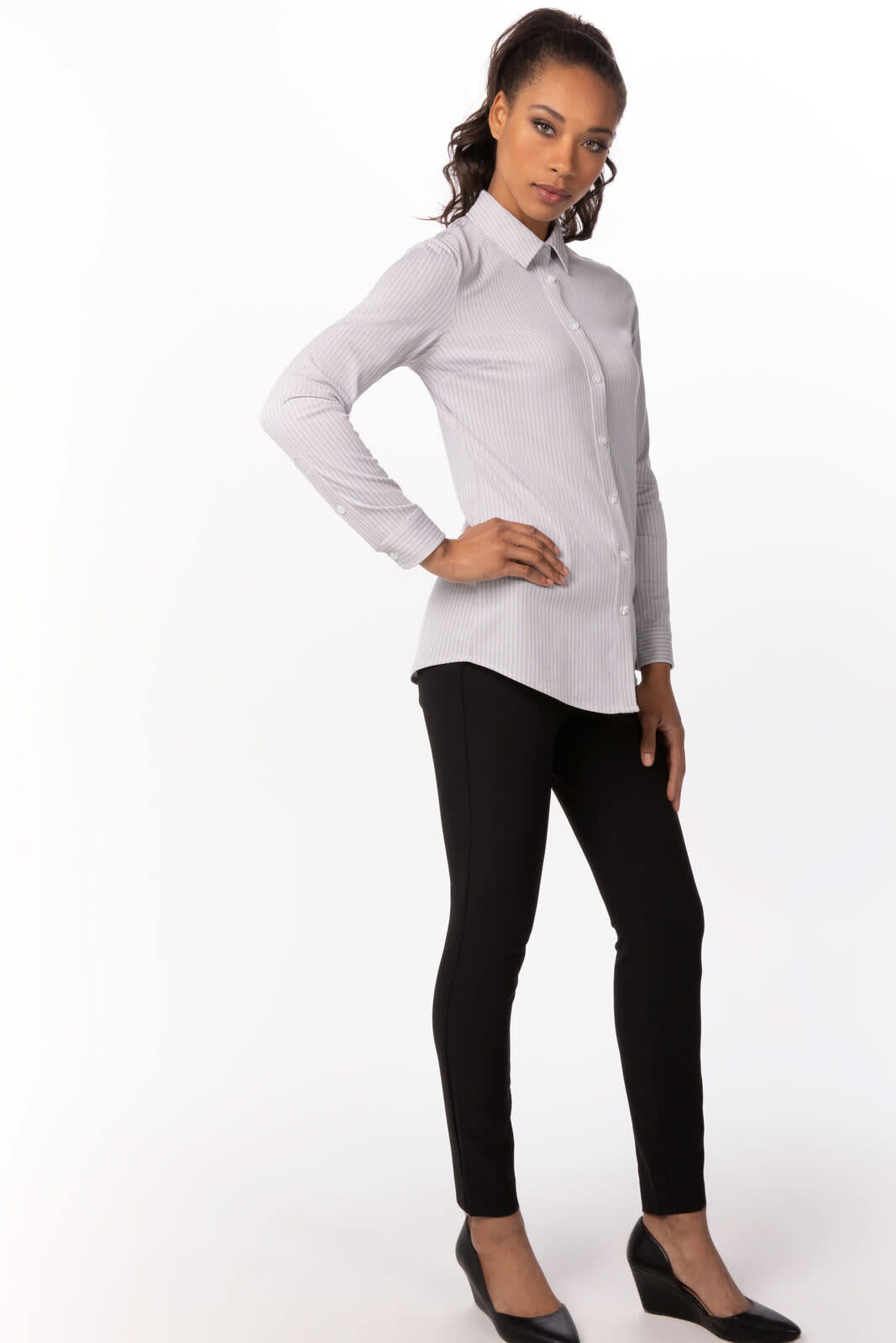 A woman stands in a light grey long-sleeved shirt paired with black trousers and shoes. The shirt features a logo.