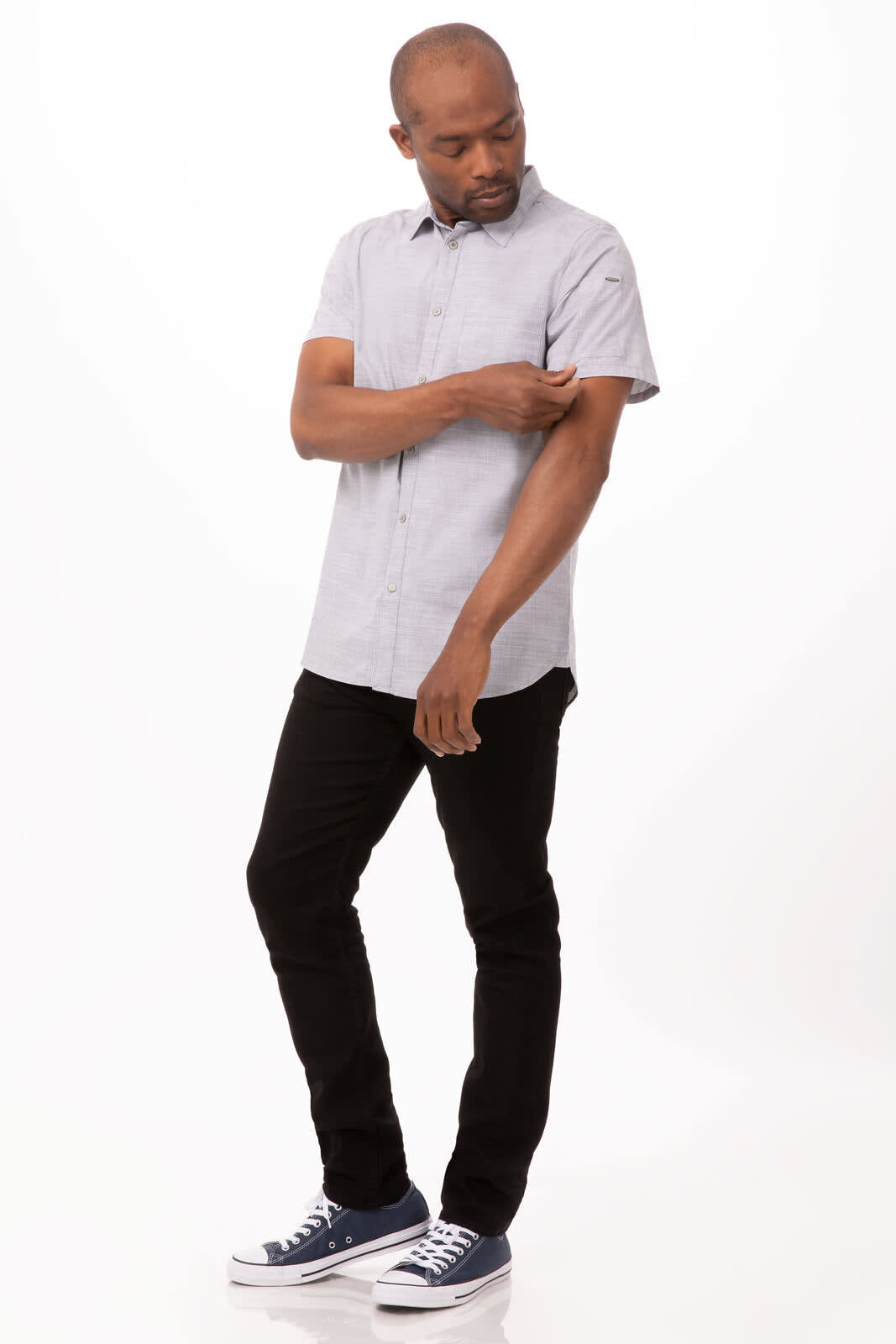 A man wearing a short-sleeved grey shirt with a collar, black trousers, and navy sneakers, adjusting his sleeve.