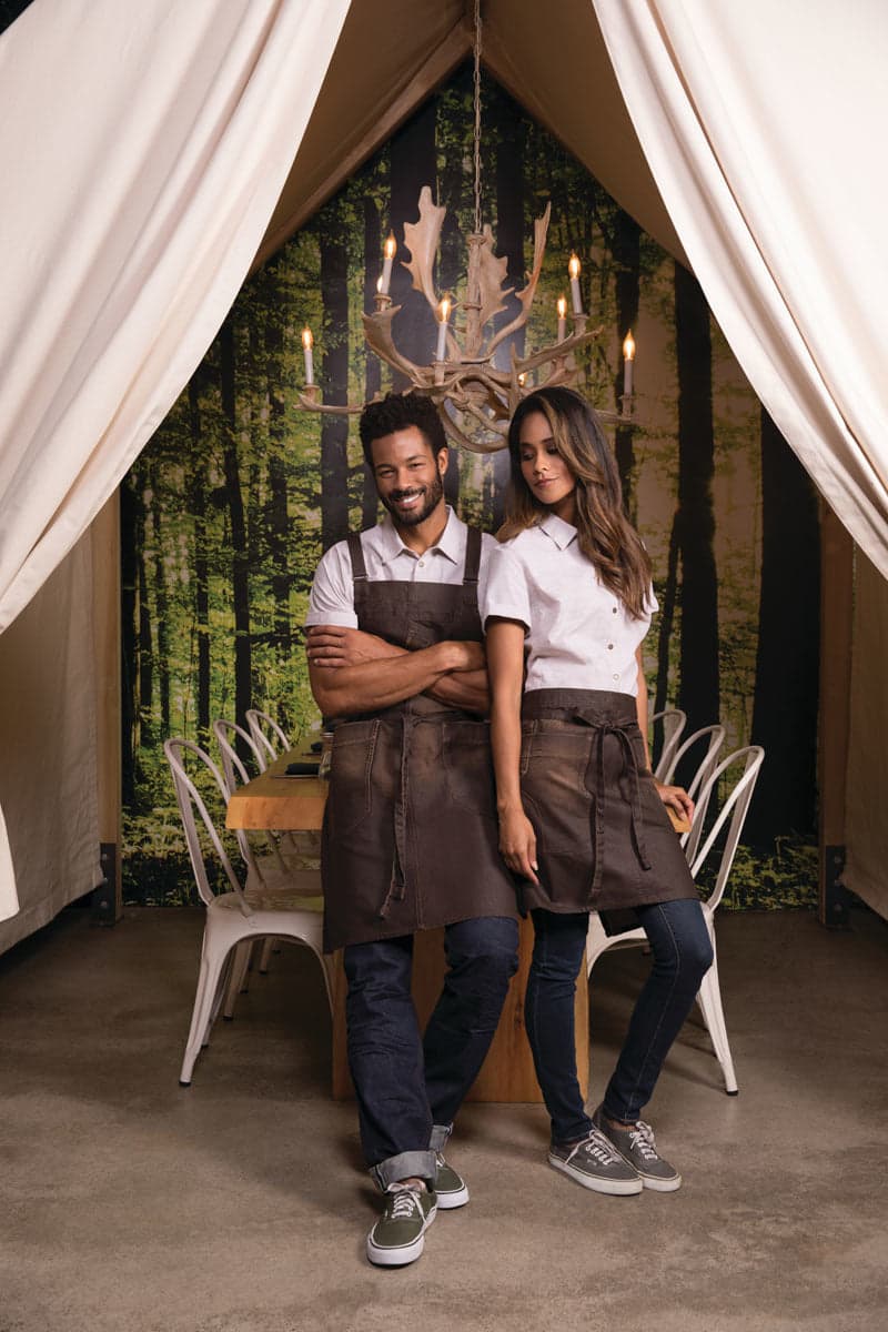 A couple stands in a forest-themed setting, wearing white shirts and brown aprons, with a logo visible on the aprons.