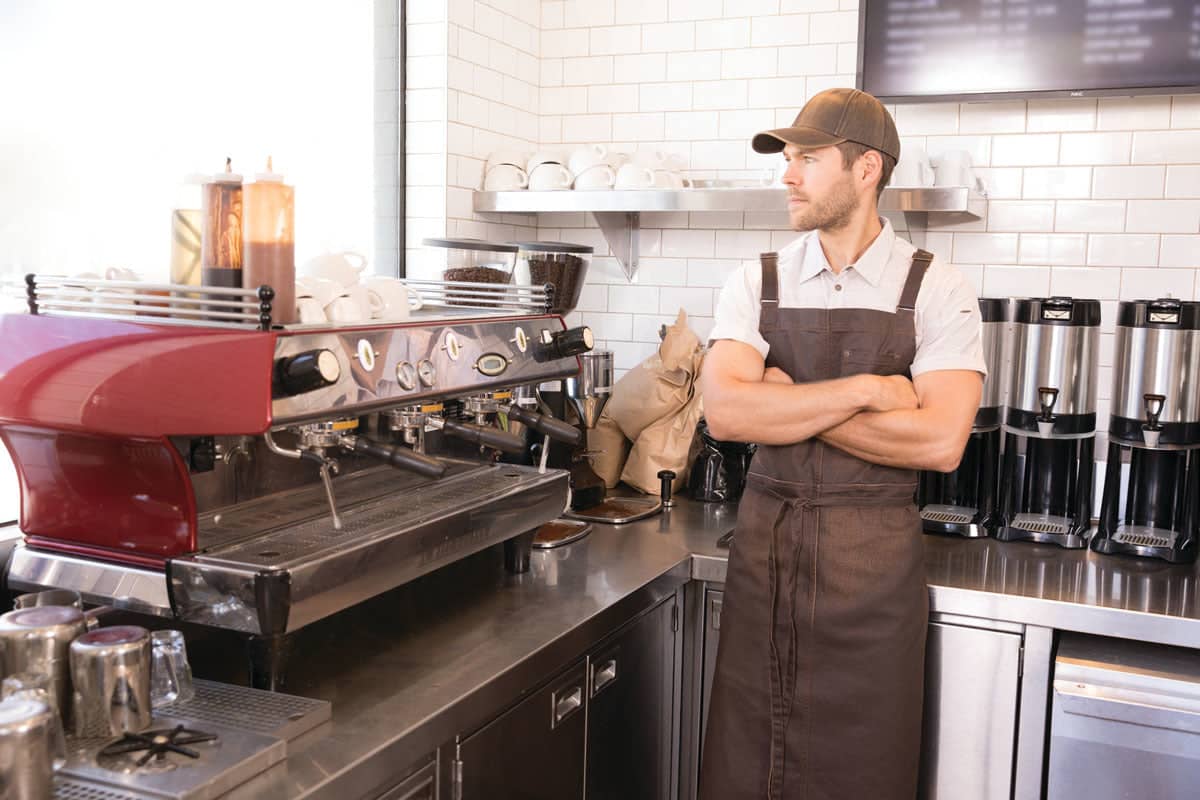 A barista in a brown apron and cap stands by an espresso machine in a coffee shop setting.