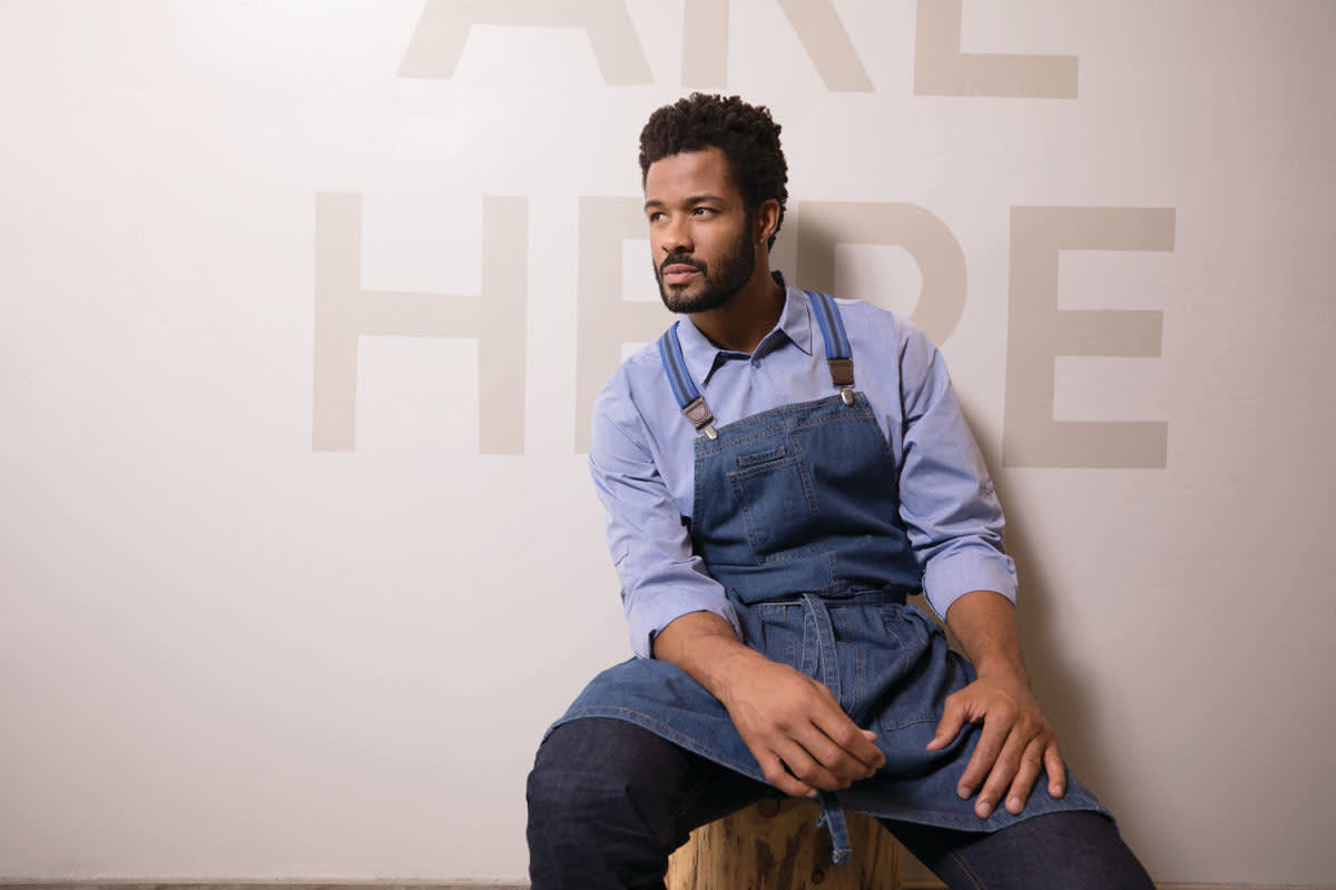 A man seated on a stool wears a blue button-up dress shirt and a matching denim apron against a light wall.