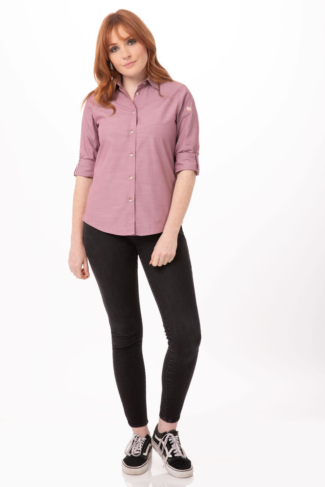 A woman in a pink chambray shirt stands against a plain background, paired with black jeans and sneakers.