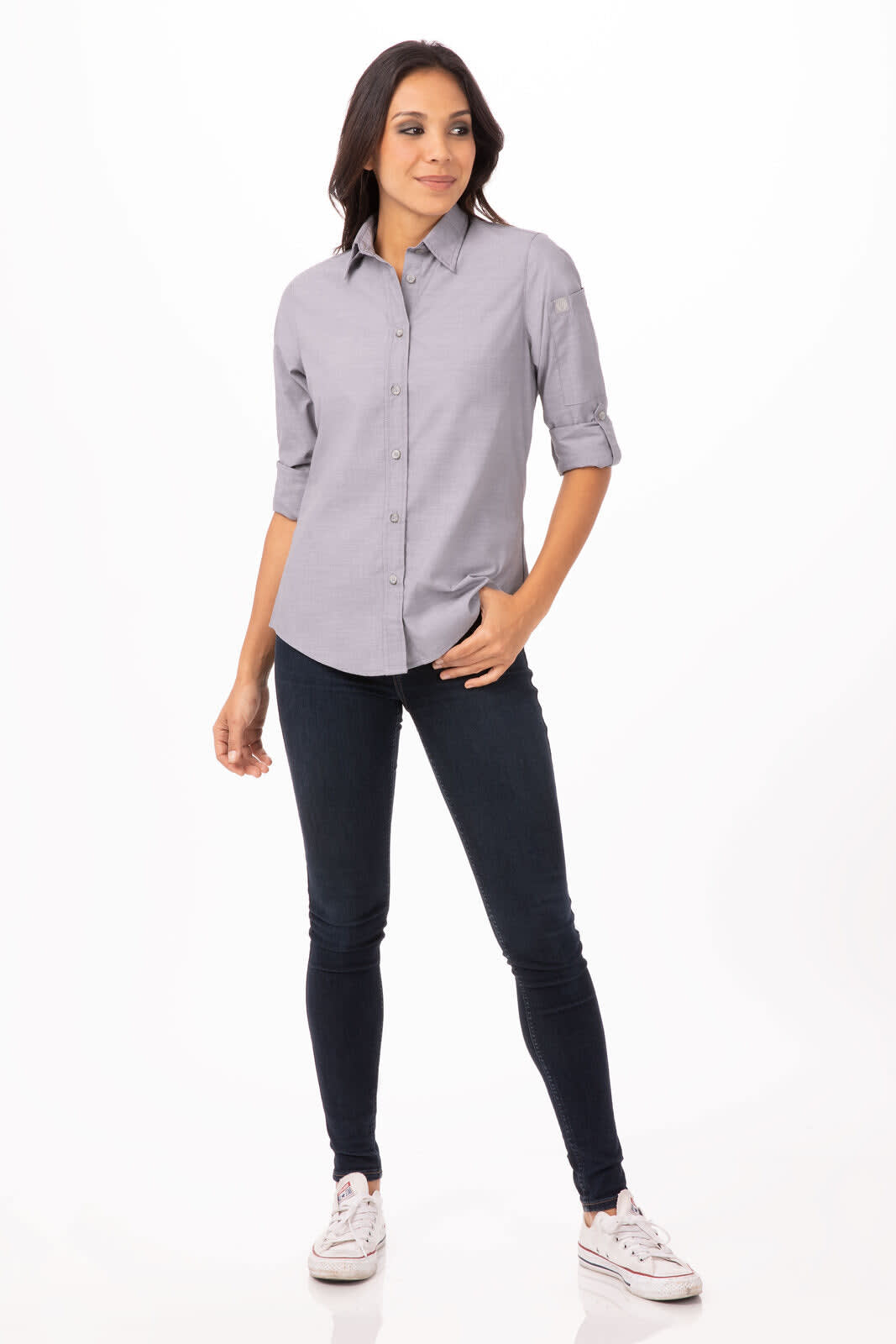 A woman stands wearing a light blue chambray shirt and dark jeans, with rolled-up sleeves and casual footwear.