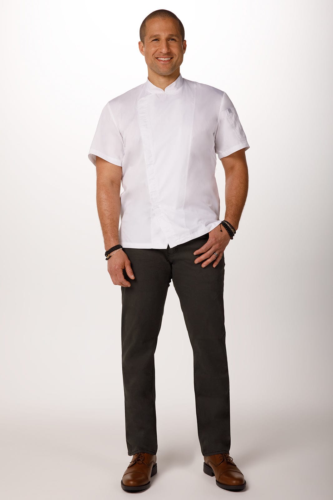 A chef jacket in white, worn by a man standing casually, paired with dark trousers and brown shoes.