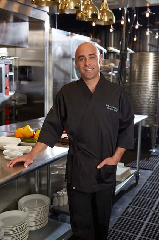 A black chef jacket worn by a smiling chef standing in a modern kitchen with various utensils and plates around.