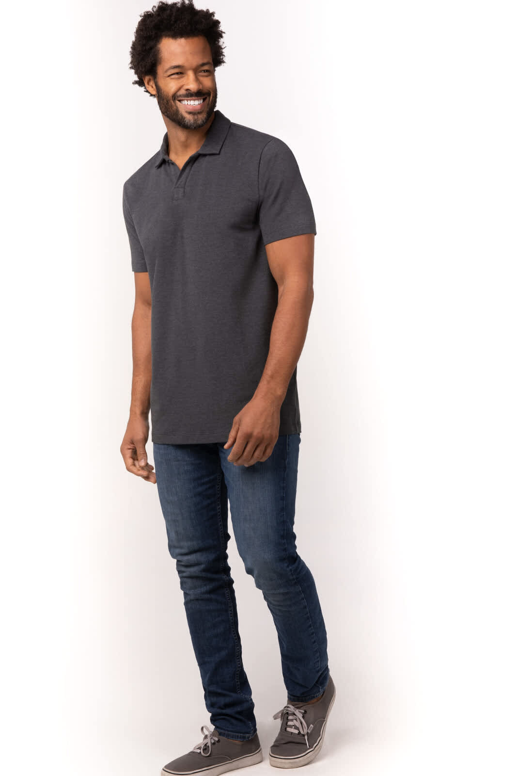 A man wearing a dark grey polo shirt and blue jeans, standing casually with a smile. The shirt has a logo.