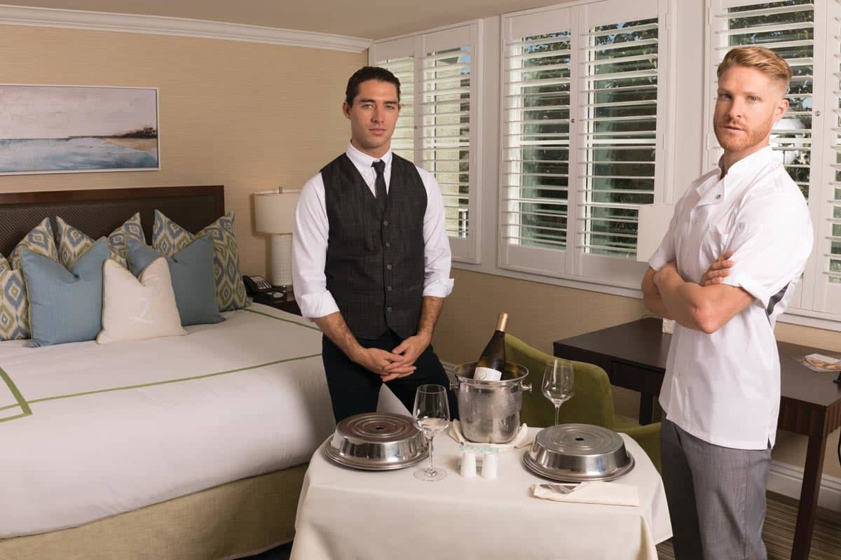 A dining setup featuring a waiter and chef beside a table with covered dishes, wine glasses, and an ice bucket in a room.