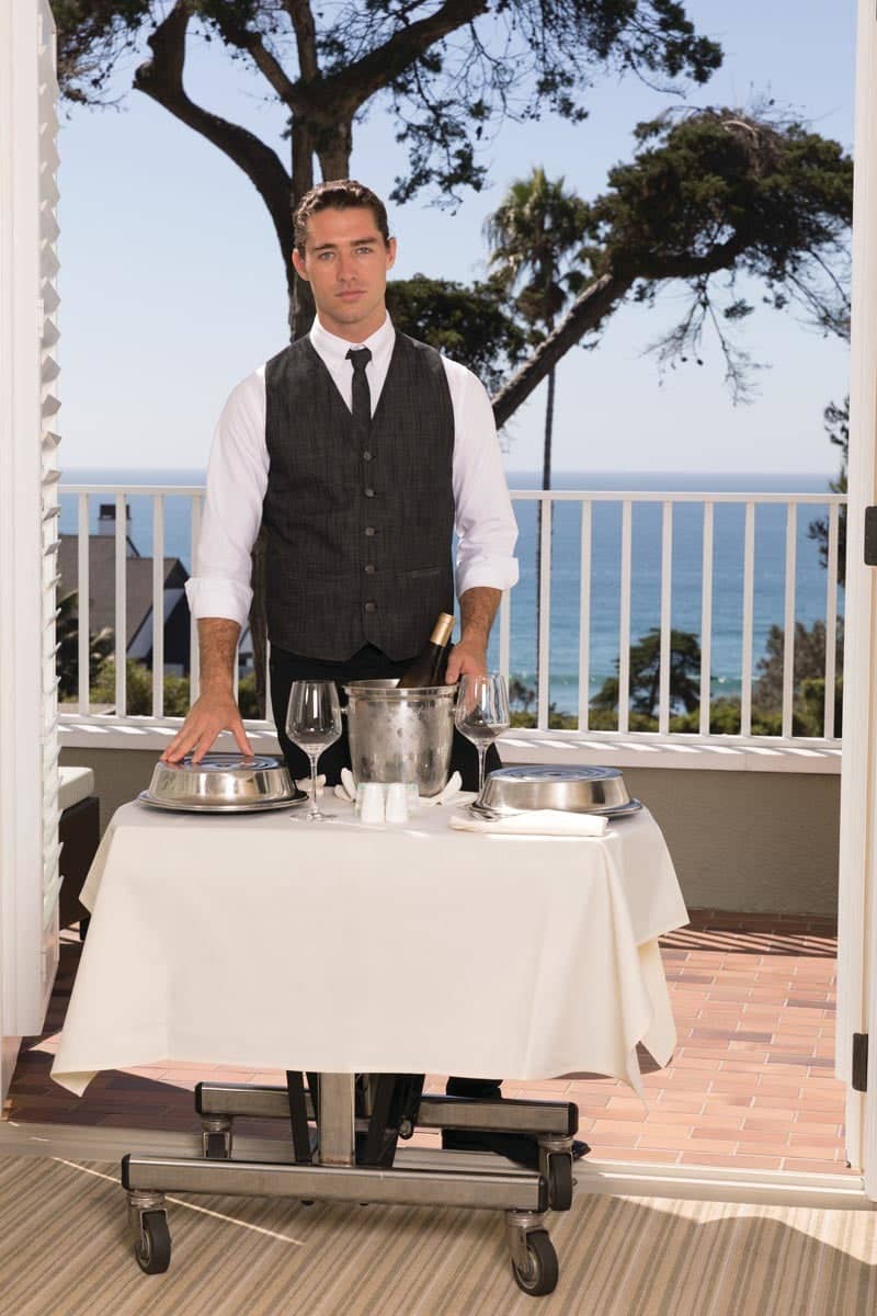 A waiter in a black vest stands beside a table with a white tablecloth, overlooking an ocean view.
