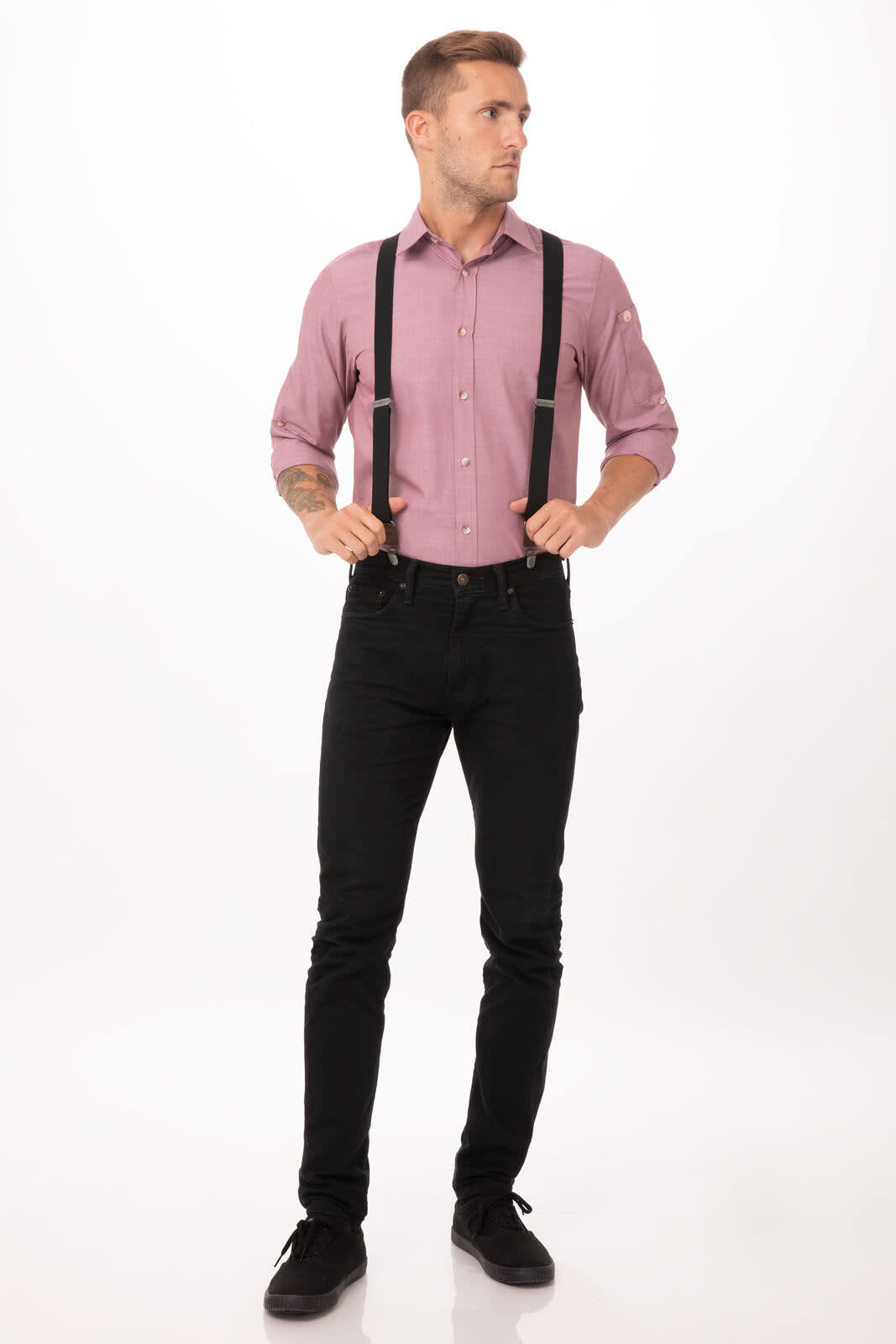 A man wearing black pant suspenders over a pink shirt and black jeans, standing against a plain background.