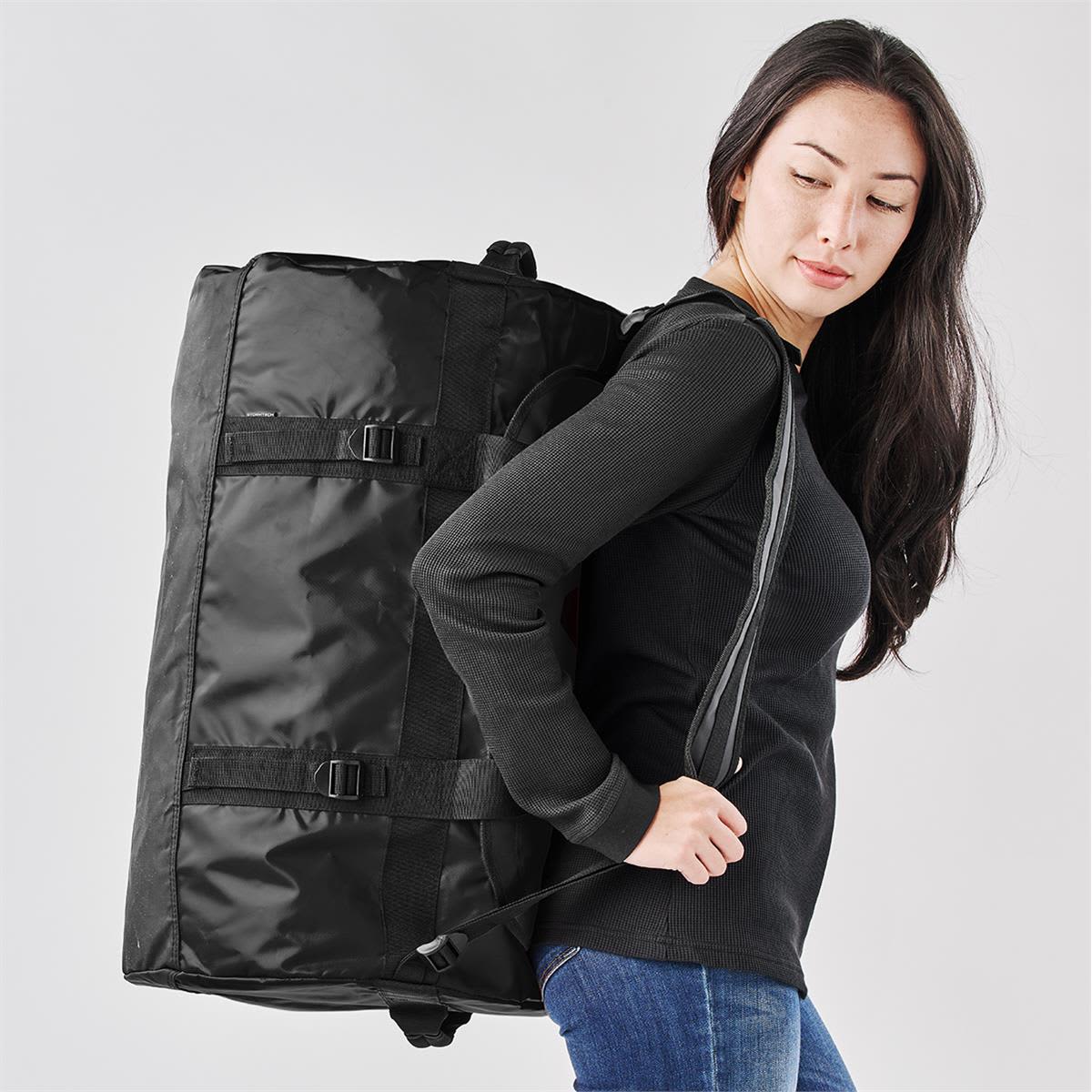 A medium black duffle bag featured being carried by a person, with adjustable straps and buckles visible.
