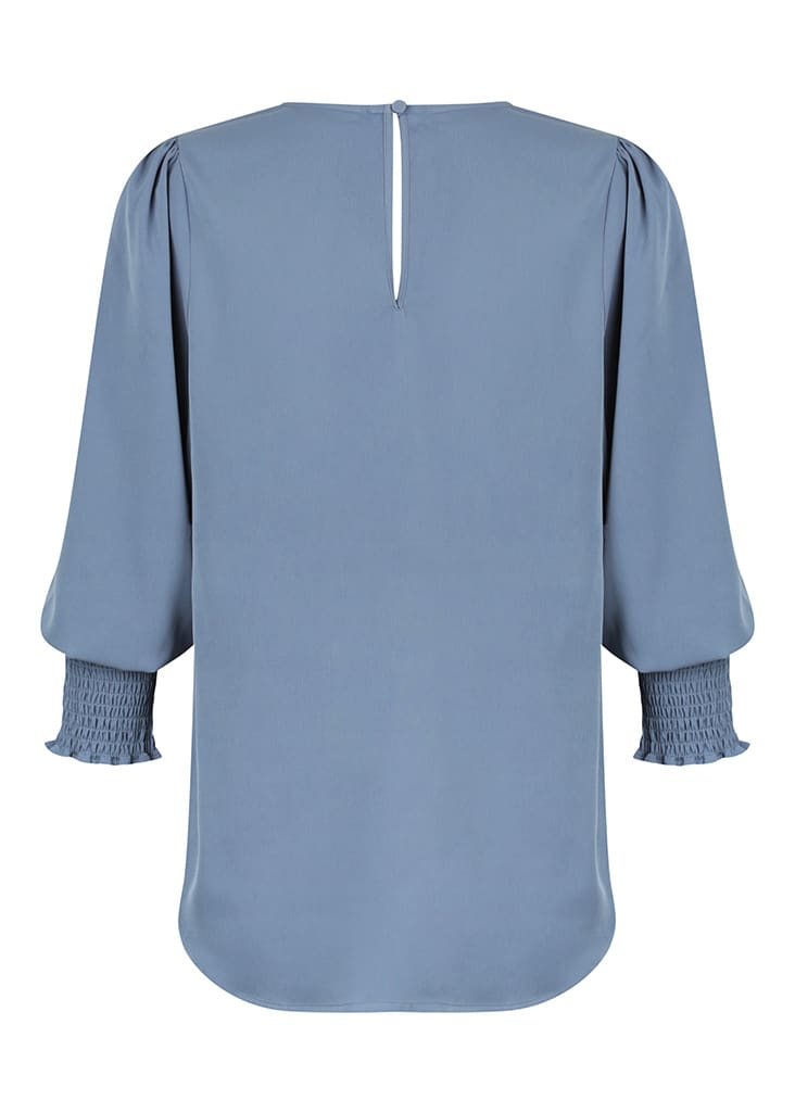 A blue blouse featuring long sleeves with smocked detail and a back keyhole opening. It has a branding logo.