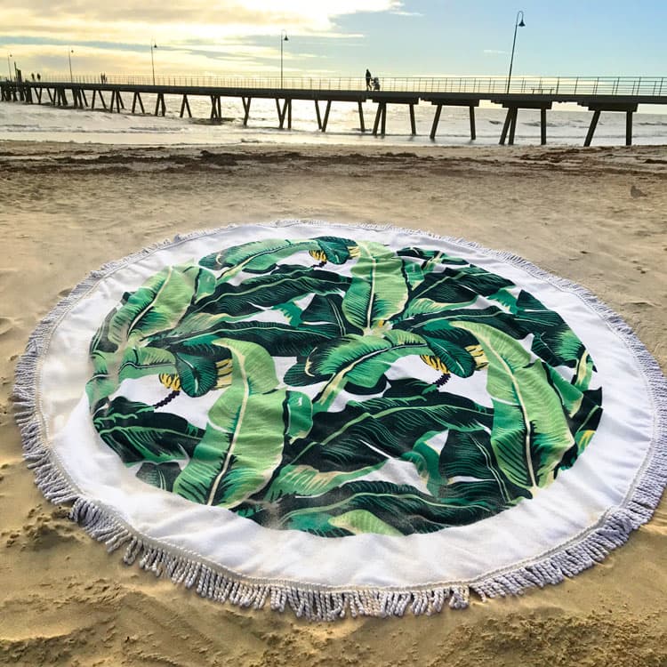 A round towel featuring green banana leaves on a white background, set against a sandy beach with a pier in the background.