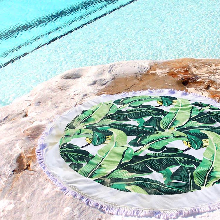 A round towel featuring vibrant green banana leaves, set on a rocky surface near a blue pool.