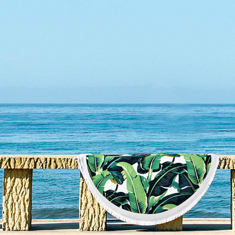 A round towel featuring a vibrant green banana leaf pattern, set against a serene beach backdrop.