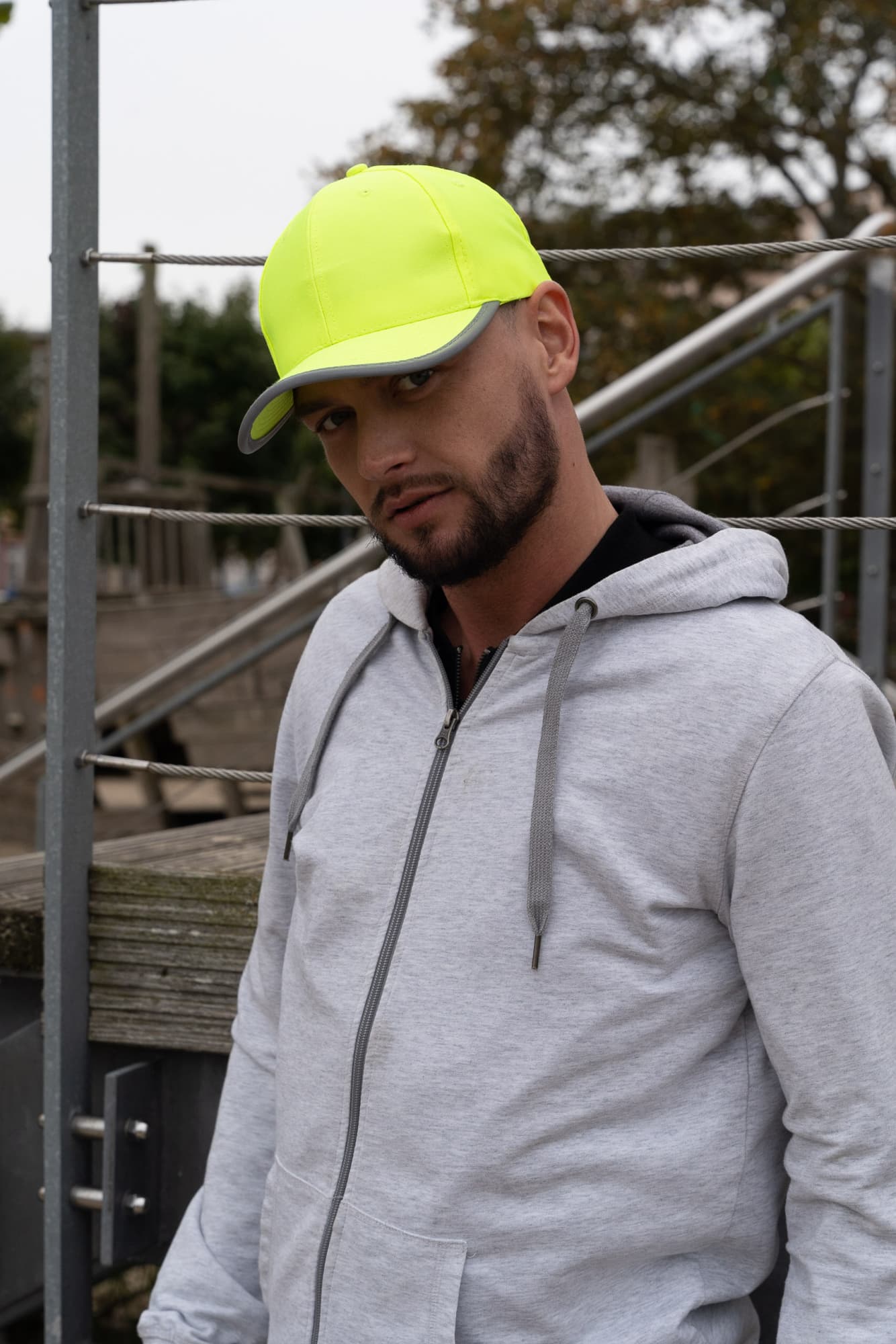 Luminescent Safety Cap with Reflective Trim HE-3021 A man wearing a bright yellow cap stands outdoors, paired with a grey hoodie. The cap features reflective trim and a logo.