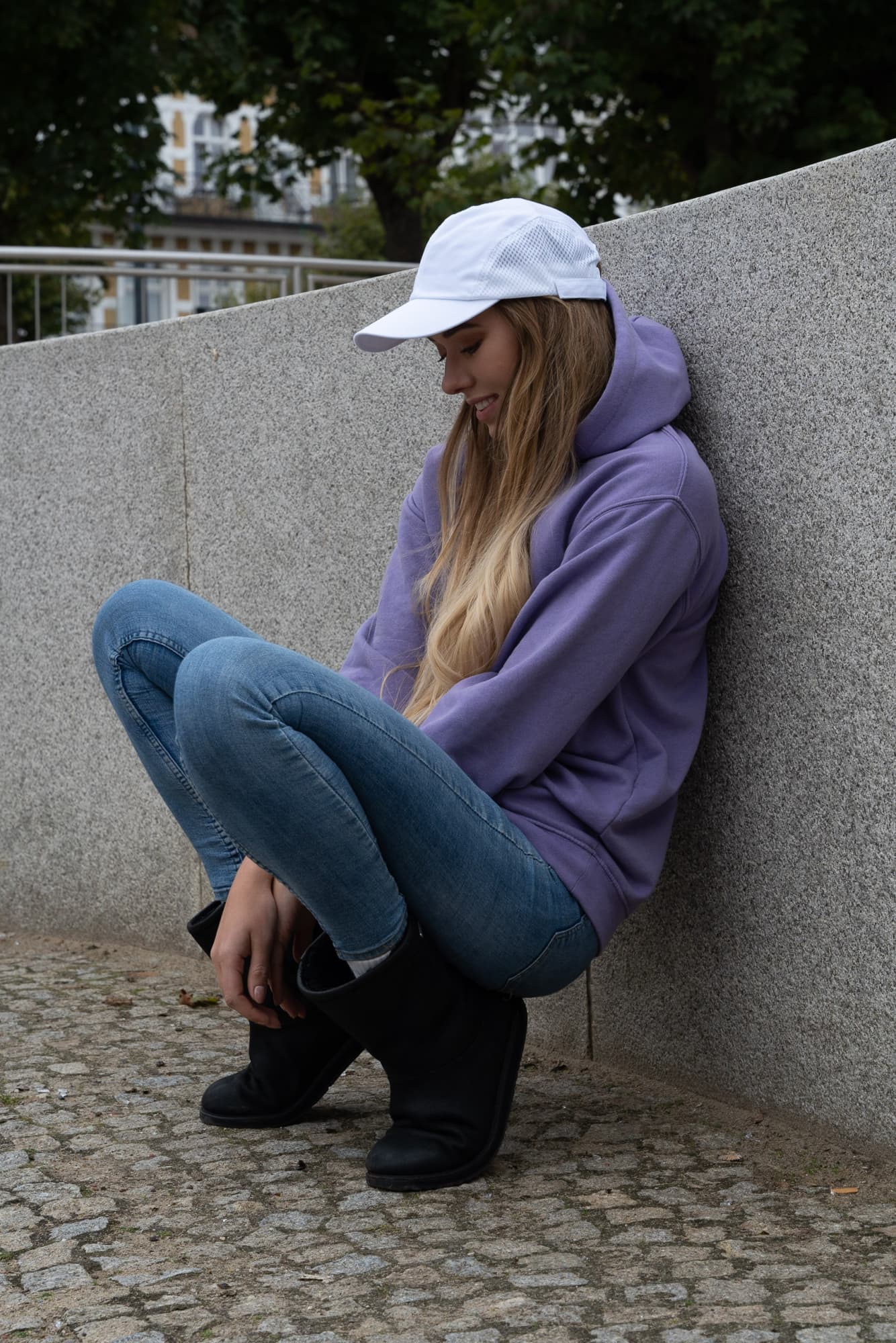Brushed Cotton HE-3812 A woman wearing a white cap sits on a grey stone surface, dressed in a purple hoodie and blue jeans.