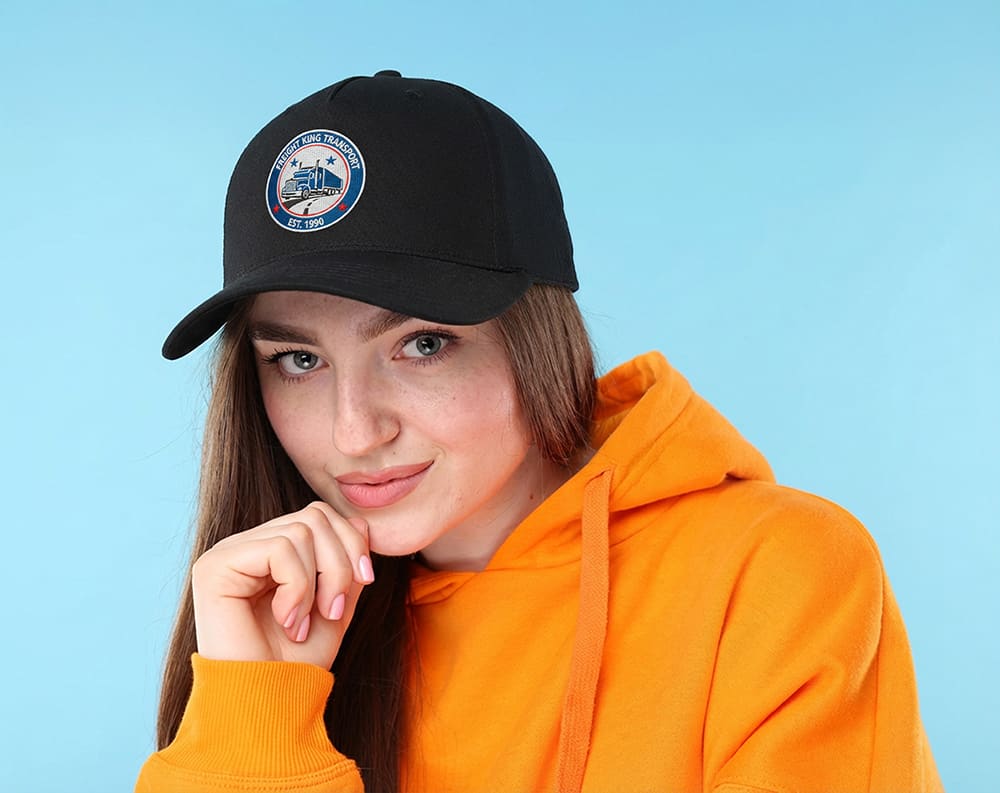 Cotton Twill Cap with Buckle HE-3963 A woman wearing a black cotton twill cap with a buckle, paired with an orange hoodie. The cap features a logo.