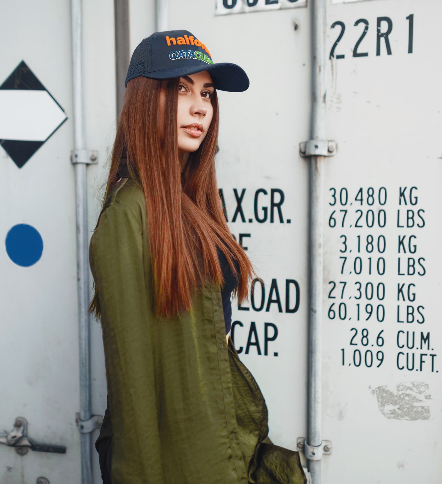 Sports Ripstop Twill with triangle perforations HE-3977 A woman with long hair wears a navy cap with a logo, standing next to a shipping container. She is dressed in an olive green jacket.