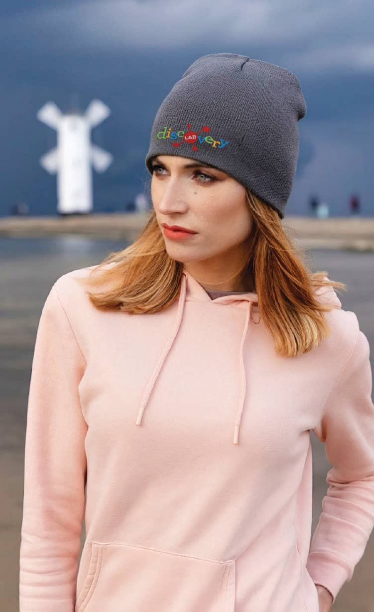 Recycled Roll Down Beanie HE-3985 A woman wearing a grey recycled roll down beanie, paired with a light pink hoodie, against a moody backdrop.