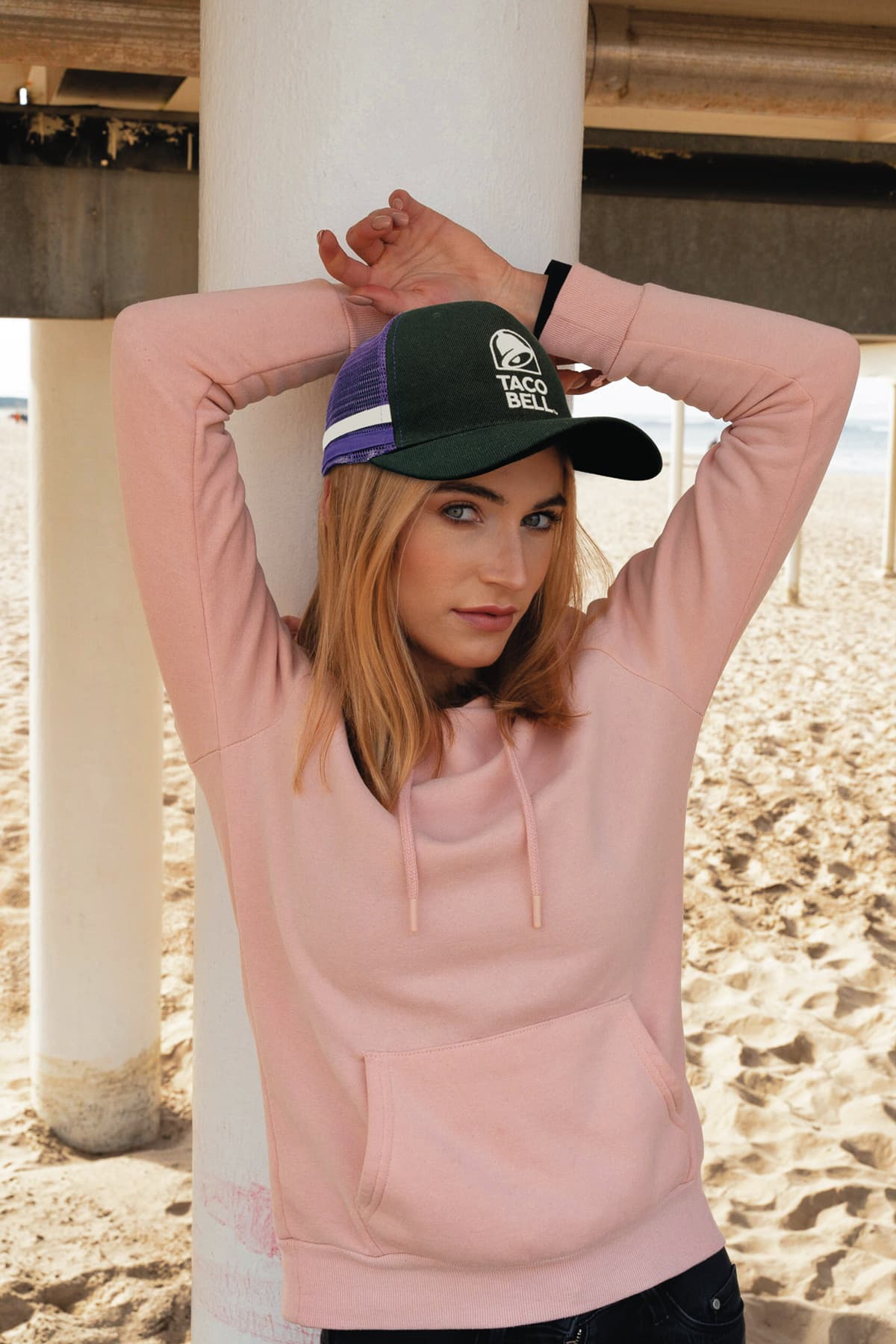 American Premium Twill/Mesh Deep Fit Trucker with Stripes HE-3995 A model wearing a pink hoodie poses under a pier, showcasing a green and purple striped trucker cap.