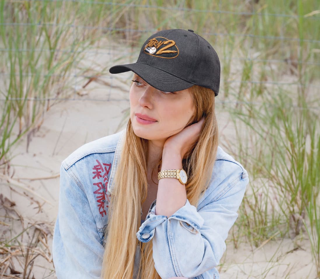 Premium American Twill Cap HE-4000 A black cap with a logo, set against a beach backdrop. The model has long hair and wears a denim jacket.