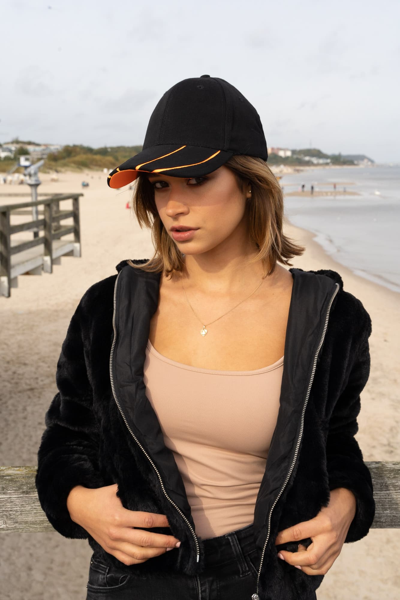 Brushed Heavy Cotton with Hi-Vis Laminated Two-Tone Peak HE-4019 A black cap with a hi-vis orange peak worn by a woman on a beach, in a casual outfit and standing near a boardwalk.