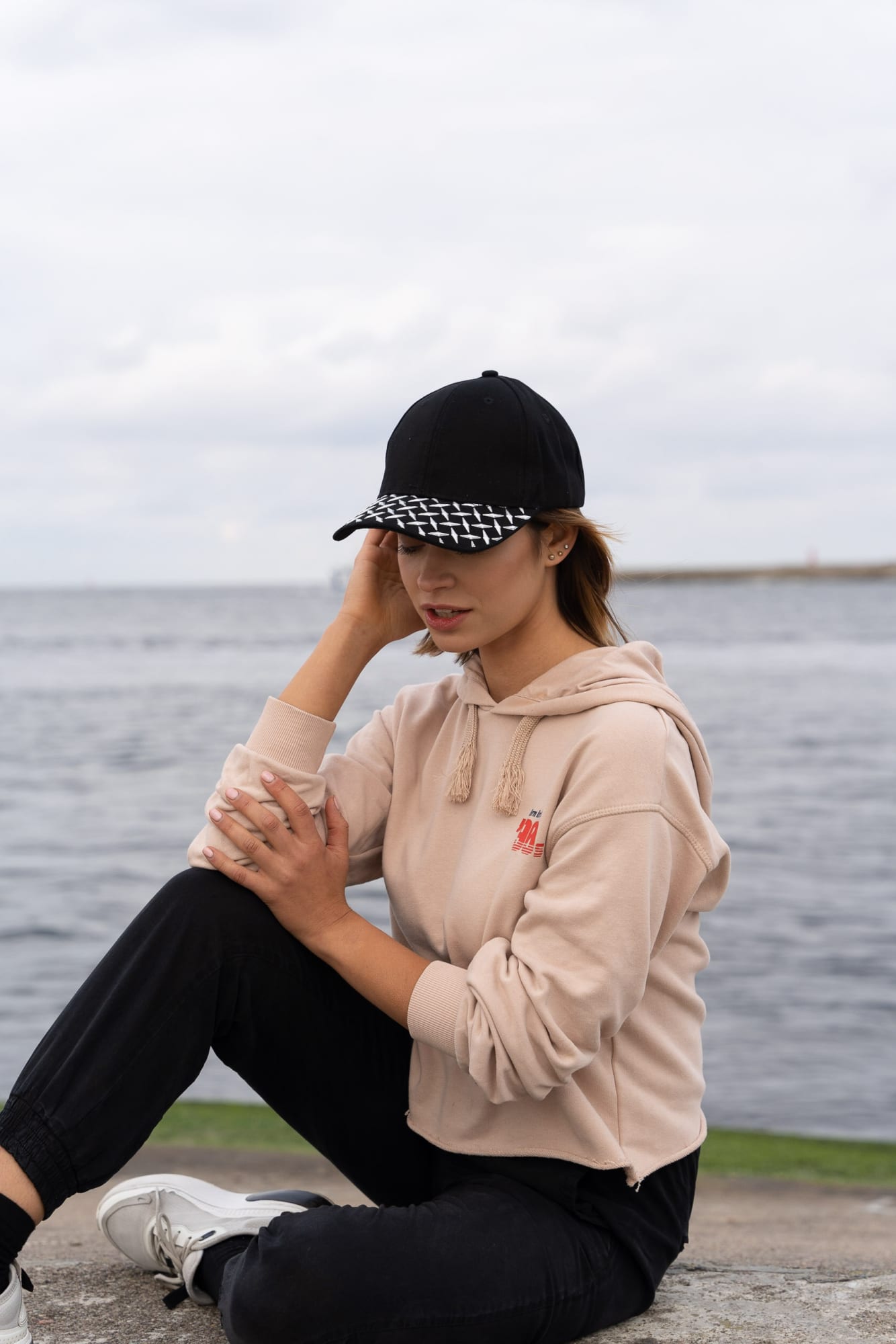 Brushed Heavy Cotton with Checker Plate on Peak HE-4044 A black cap with a checker plate peak, worn by a woman in a beige hoodie, with a scenic water background.