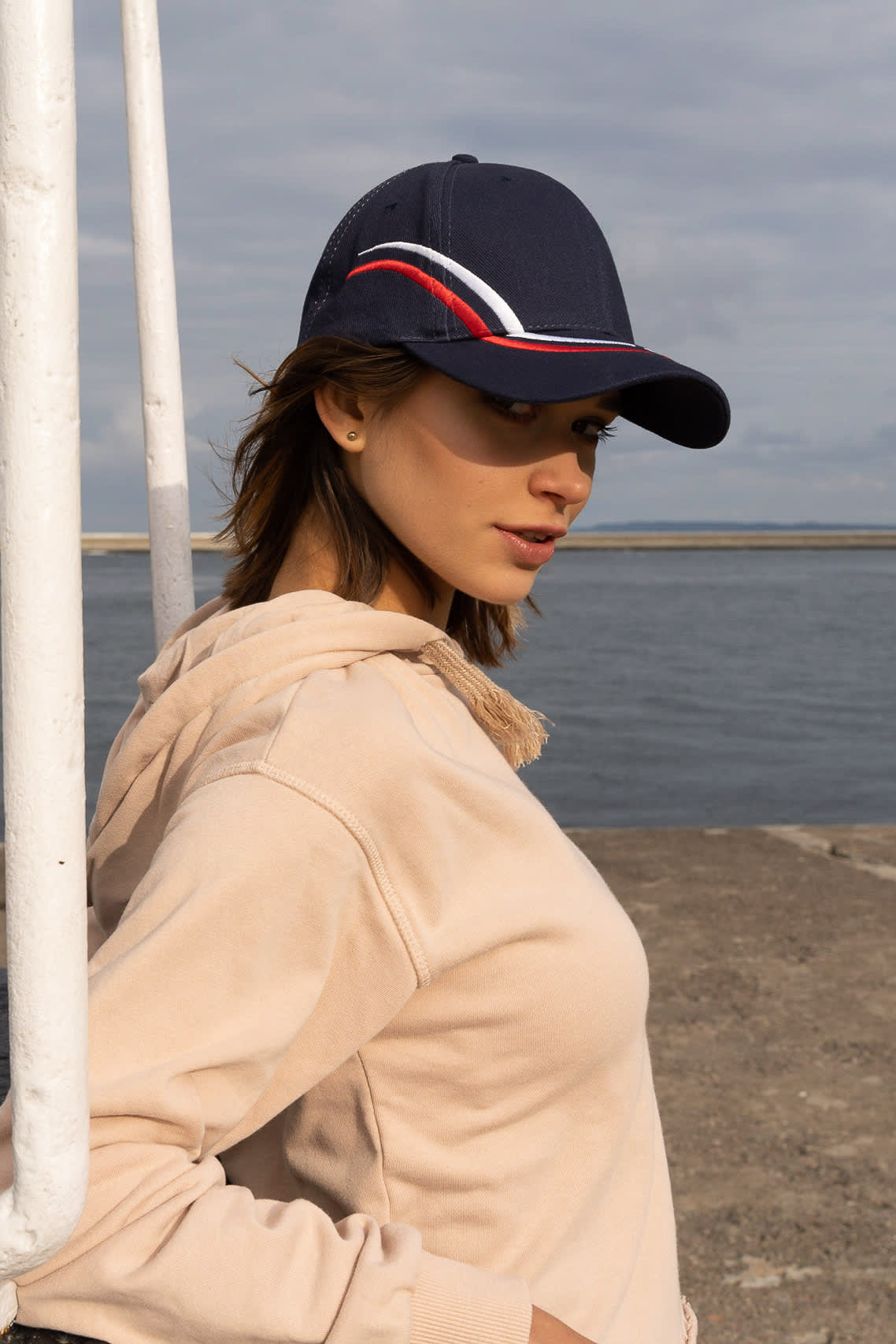 Brushed Heavy Cotton with Curved Embroidery on Crown and Peak HE-4075 A navy cap with curved embroidery in red and white, worn by a woman near a waterfront.