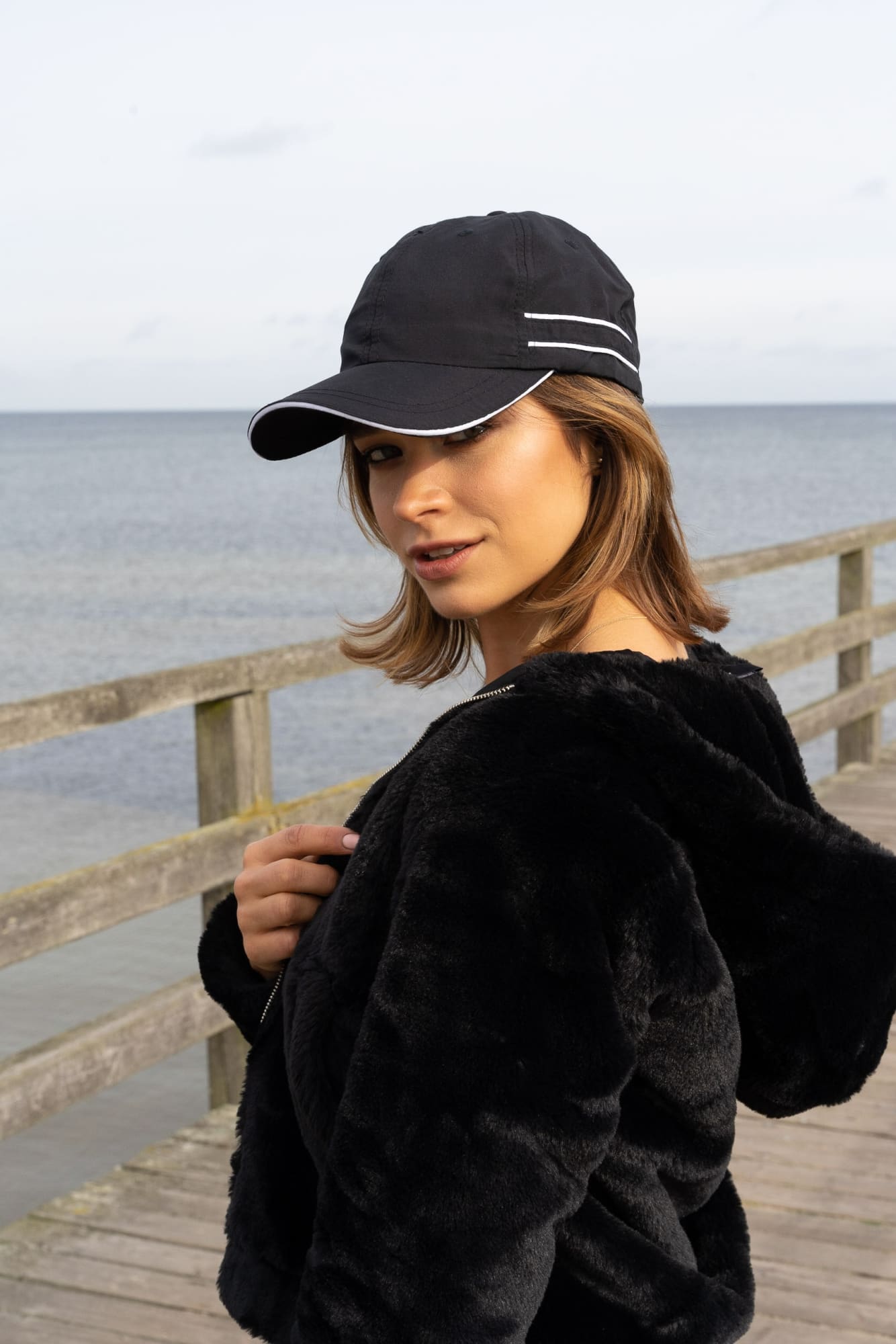 Microfibre Sports Cap with Piping and Sandwich HE-4077 A black microfibre sports cap with white piping, worn by a woman by the seaside. The setting includes a wooden railing.