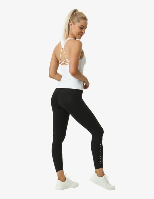 A woman in a white tank top and black leggings performs a yoga pose with her arms raised. The leggings feature a logo.