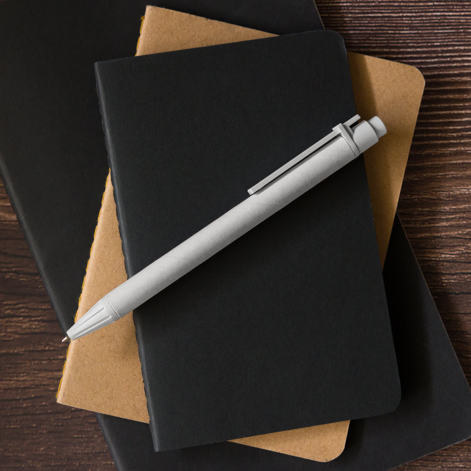 A grey pen rests on top of two notebooks, one black and one brown, on a wooden surface.