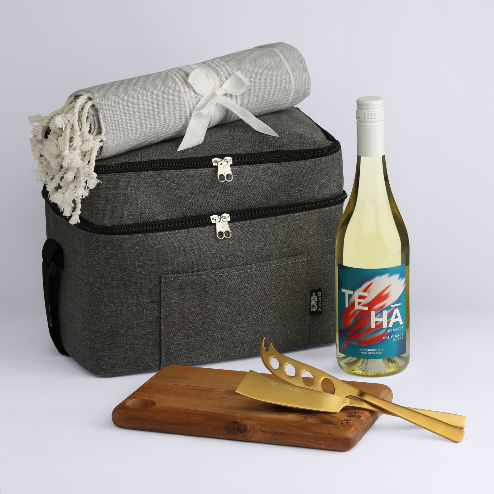 A cooler bag in grey with a blanket, a bottle of white wine, a wooden cutting board, and gold utensils.