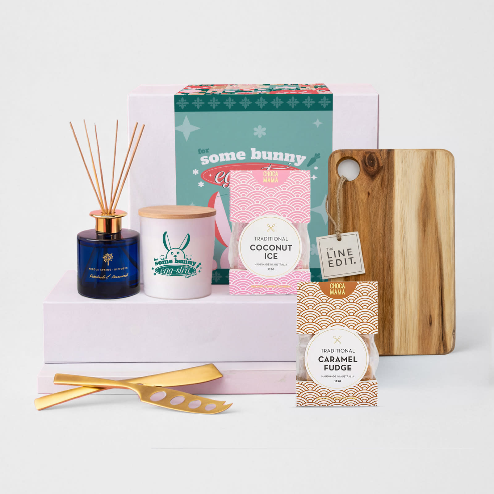 A wooden cheese board, two dessert packets, a candle, a reed diffuser, and golden serving tools, all in a gift box.