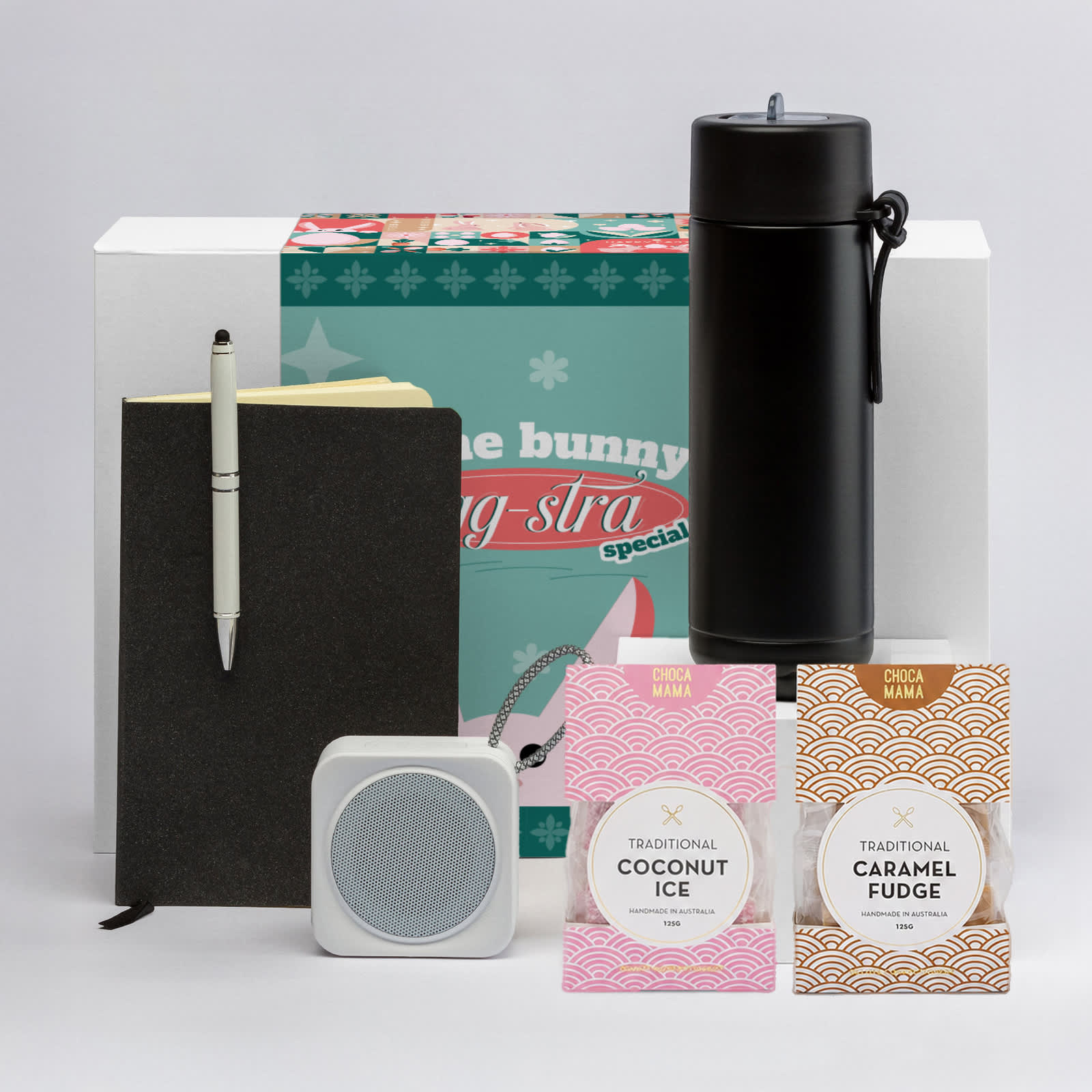 A black drink bottle, notebook, pen, wireless speaker, and two boxes of caramel fudge and coconut ice.