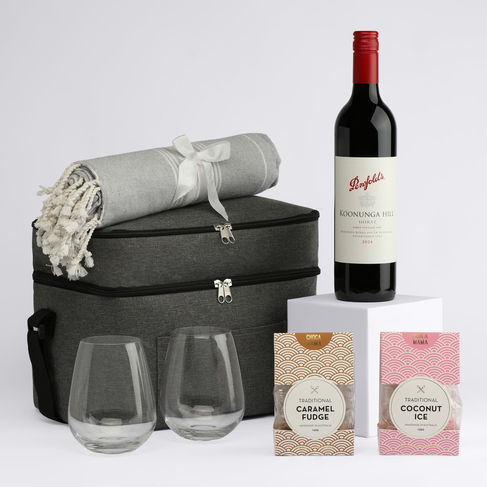 A drink bottle of red wine with two clear glasses, a grey blanket, and two packets of sweets in a stylish bag.