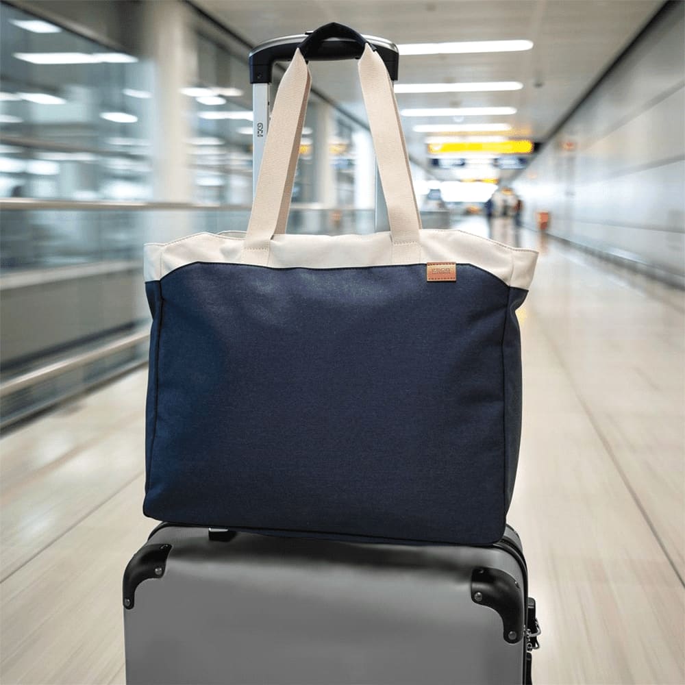 A navy blue tote bag with cream handles positioned on top of a suitcase in an airport setting.