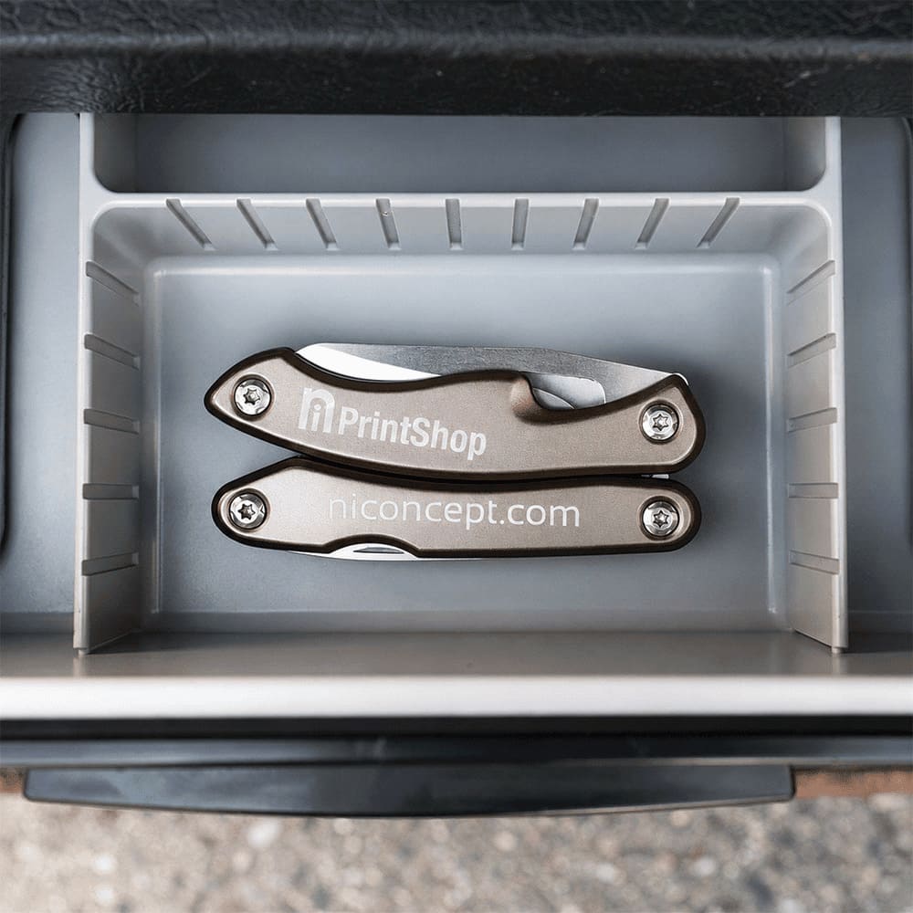 A silver multi-tool featuring several tools, placed inside a grey storage compartment. The tool has a branded logo.